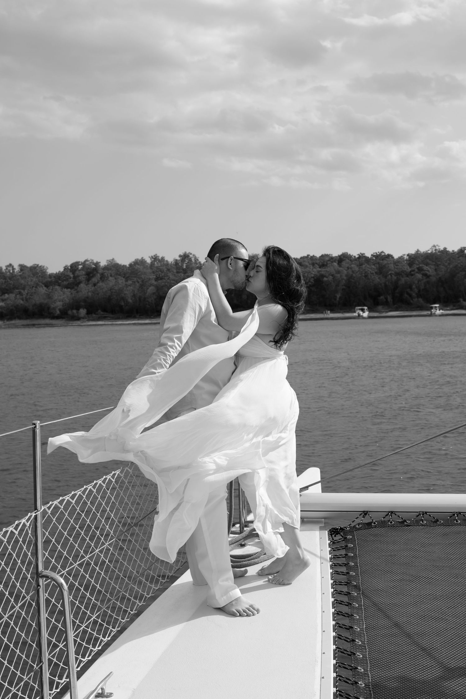 Couple kissing on sailboat bow in black and white, bride in flowing white dress, groom in white suit, water background.