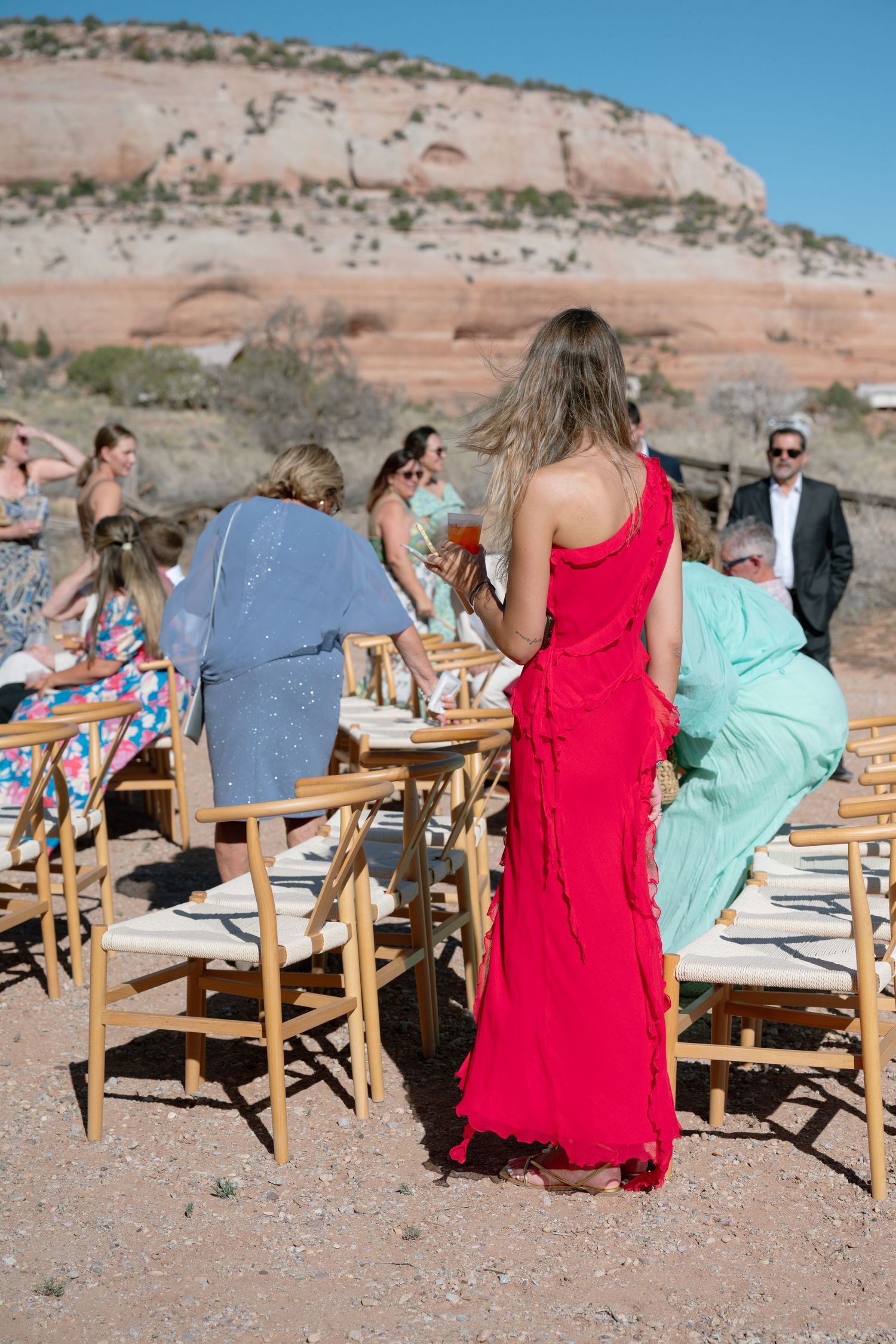 Wedding guest in elegant red dress stands among seated guests at outdoor desert ceremony with red rock cliffs backdrop.