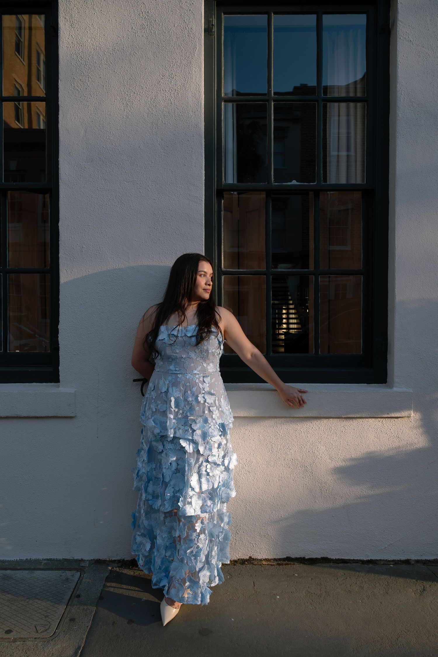Woman in blue tiered sequin gown posing against white building wall in golden hour sunlight.