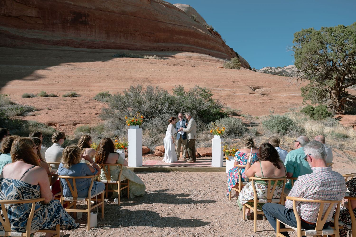 Outdoor desert wedding ceremony with guests seated on wooden chairs amid red rock formations under a clear blue sky.