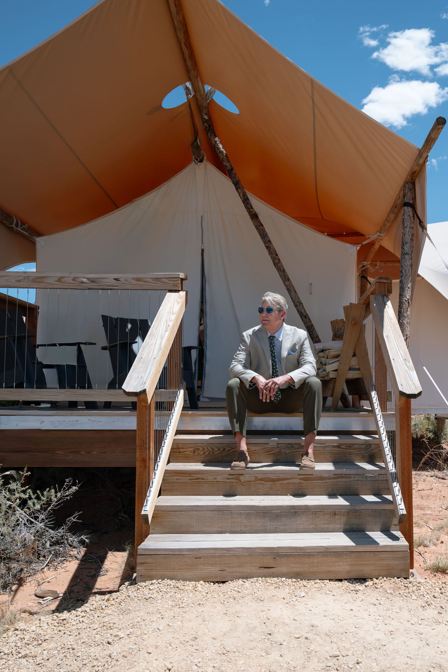Man in suit sitting on wooden steps of a luxury glamping tent in a desert landscape under a blue sky.