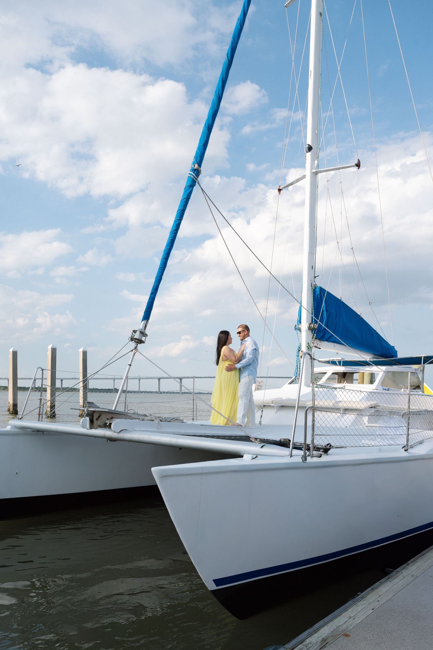 Couple in yellow dress and white outfit embracing on a white catamaran sailboat docked at a marina under cloudy skies.