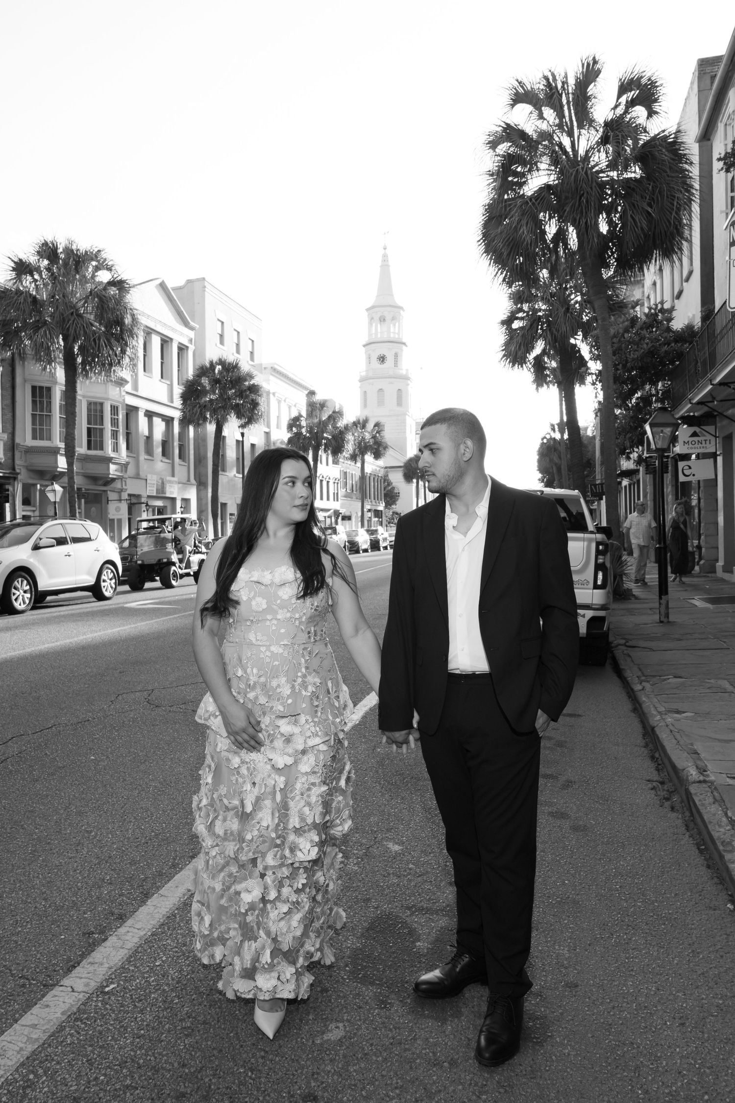 Black and white photo of an elegant couple holding hands and walking down a historic Charleston street with palm trees.