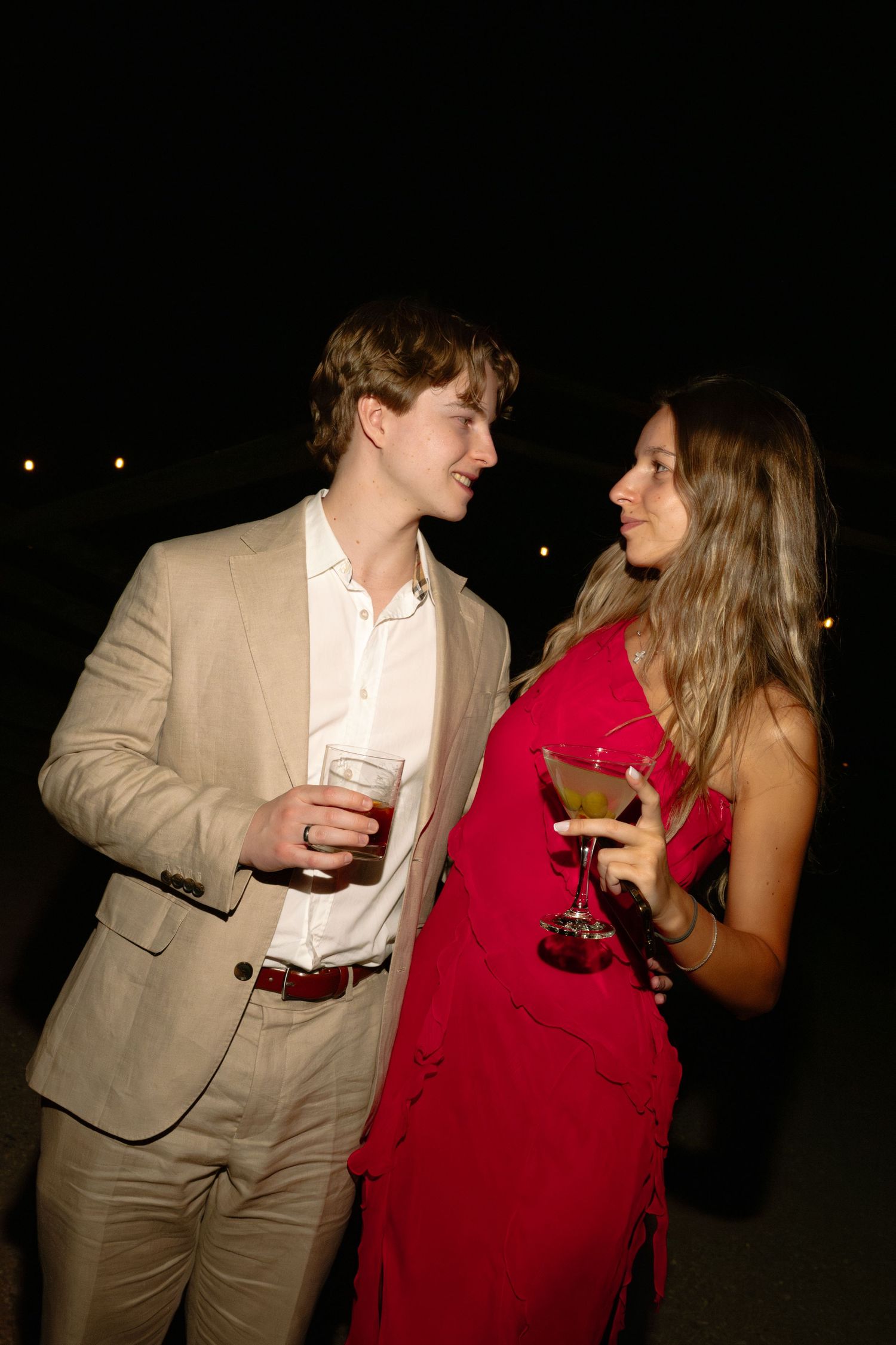 A young couple in formal attire share a romantic moment while holding cocktails at a nighttime outdoor event.