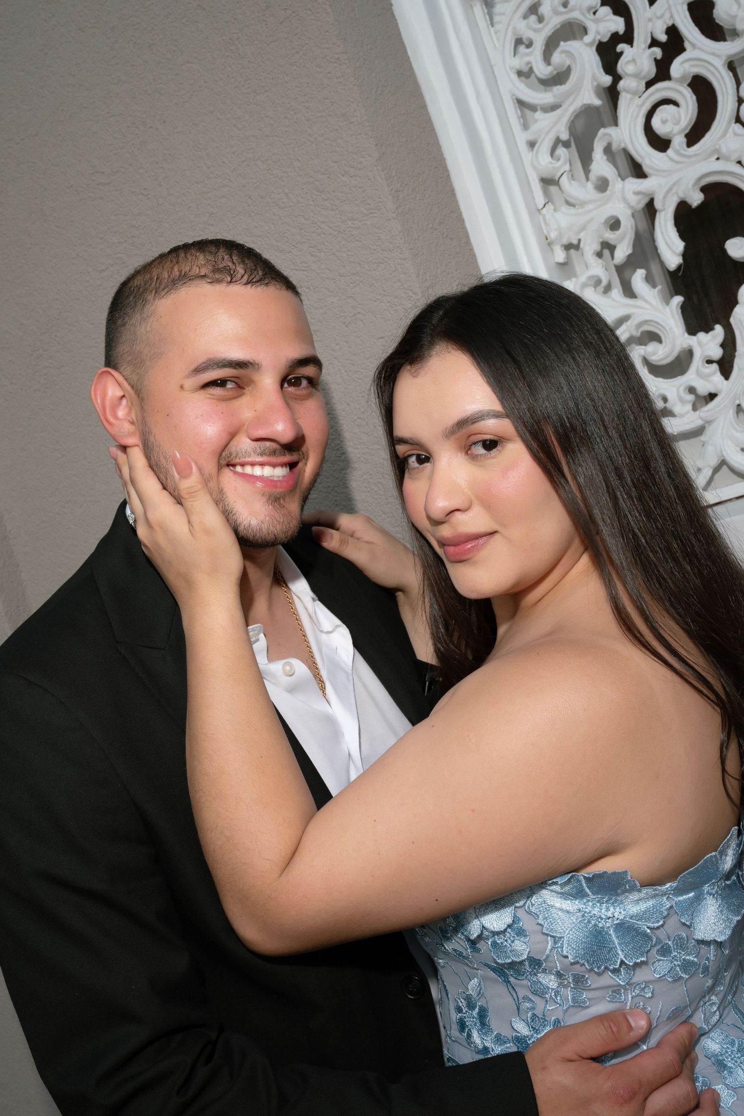 A smiling couple poses elegantly, man in black suit, woman in blue lace dress, at a formal event.