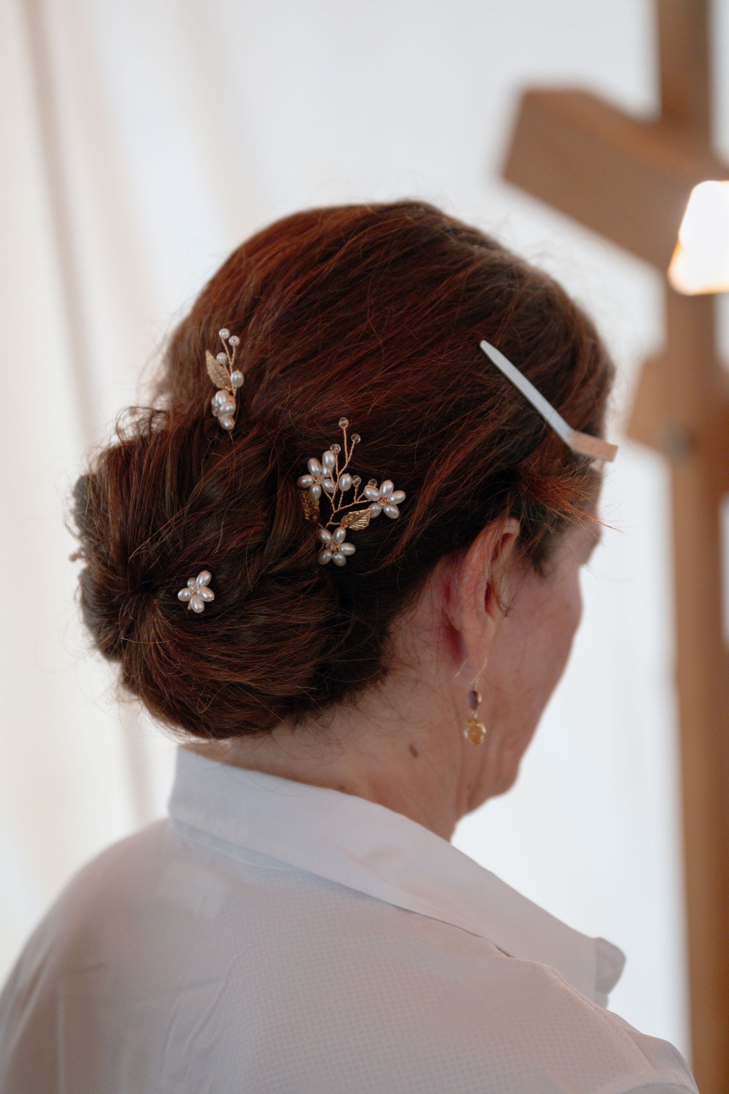 Woman with auburn hair in elegant updo adorned with delicate pearl and gold floral hair pins, wearing white top.