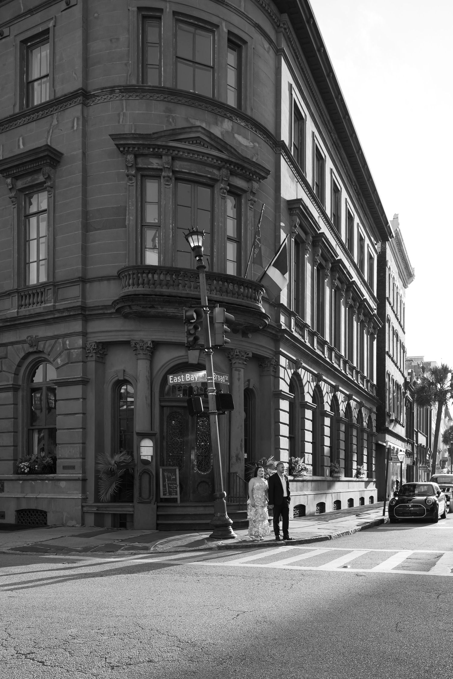 Black and white photo of a couple in wedding attire standing at a street corner beside a historic ornate building.