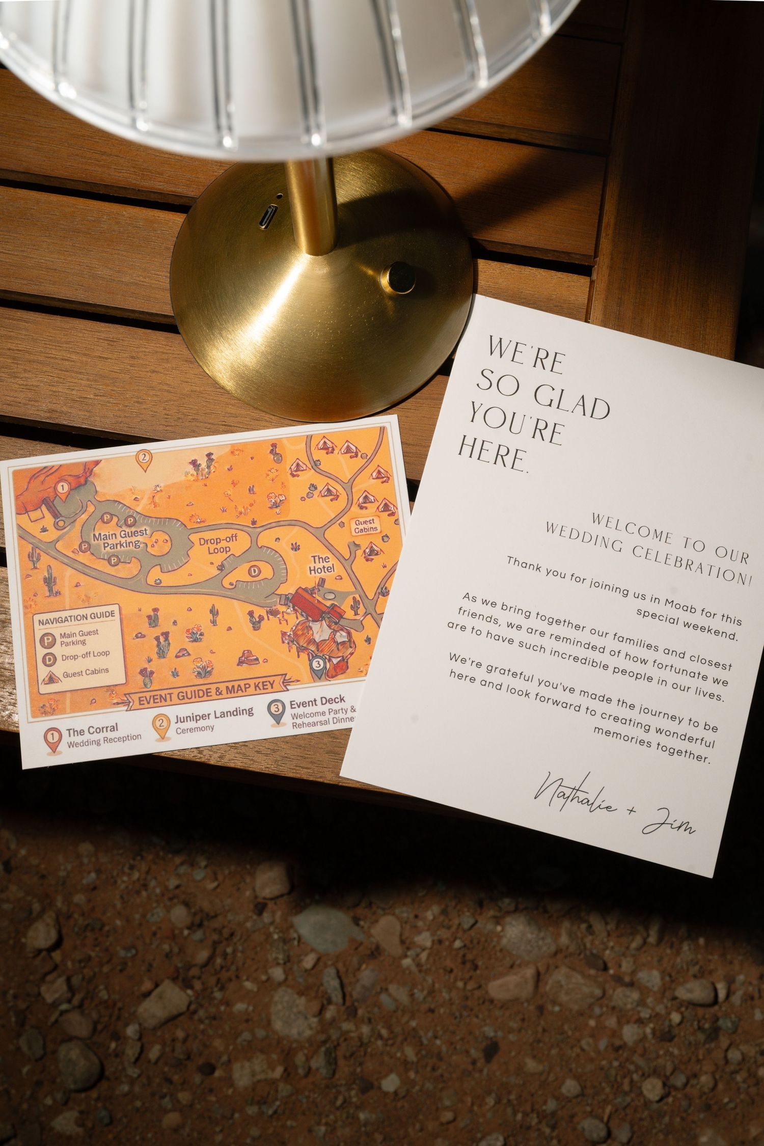 Wedding welcome card and illustrated venue map displayed on wooden table with gold lamp at elegant celebration.