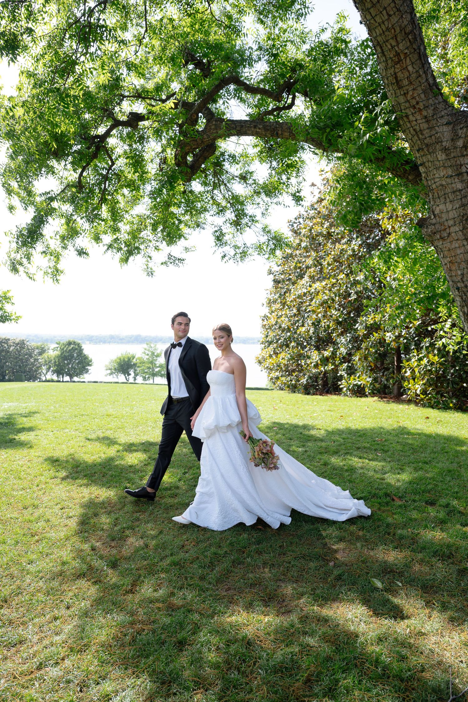 Bride in white gown and groom in black tuxedo pose under a large oak tree on a green lawn near a waterfront.
