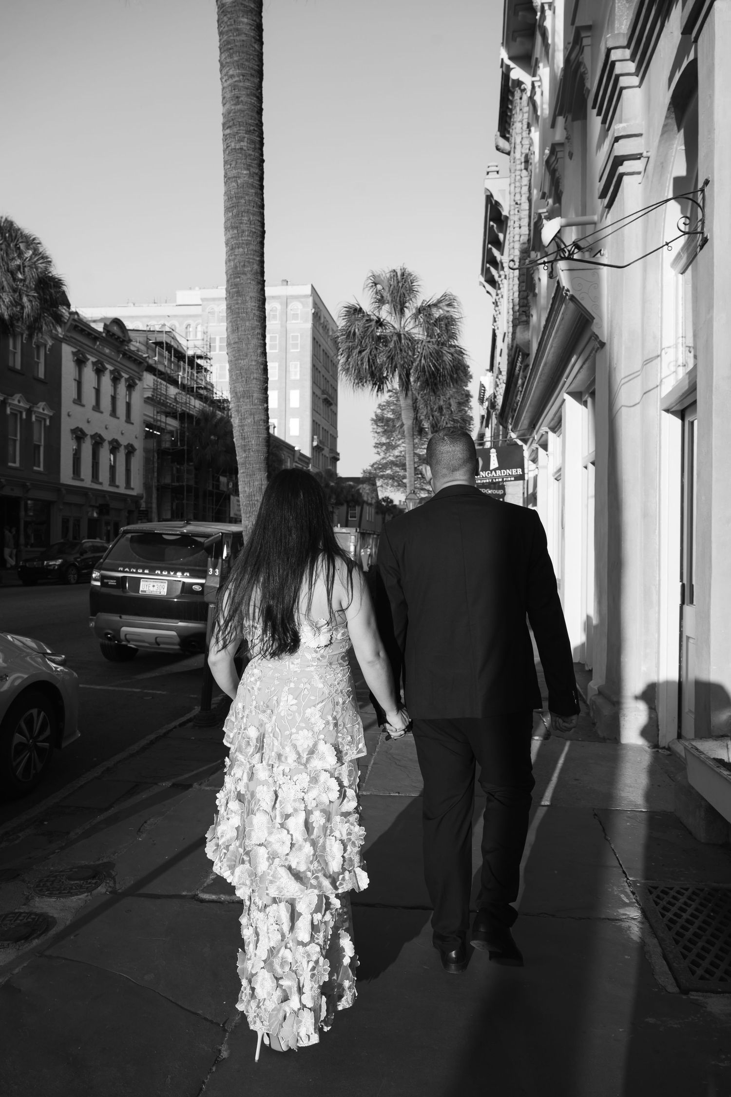 Black and white photo of a couple in formal attire walking hand in hand on a city sidewalk lined with palm trees.