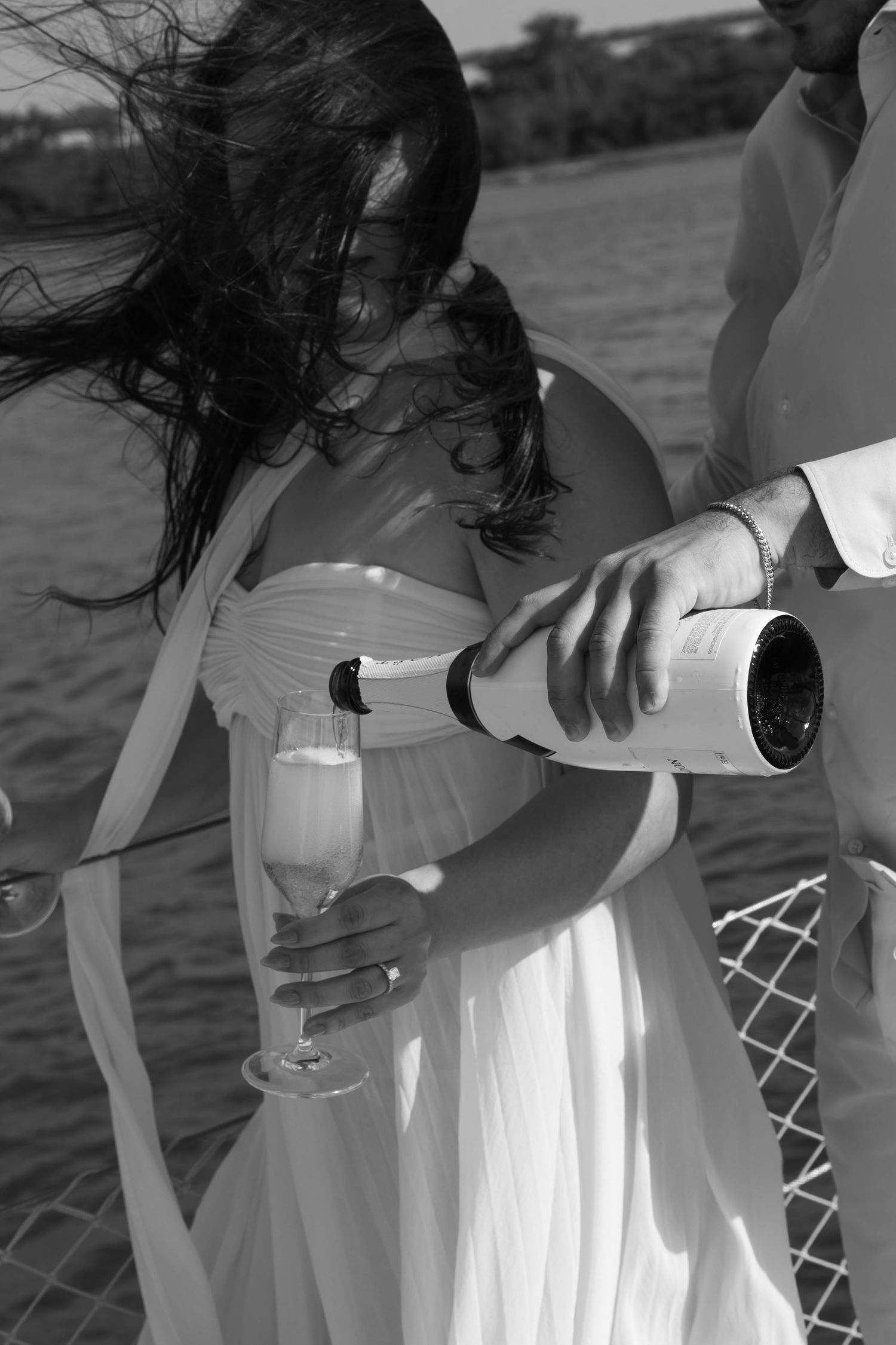 Black and white photo of a couple on a boat, man pouring champagne into a glass held by a woman in a white dress.