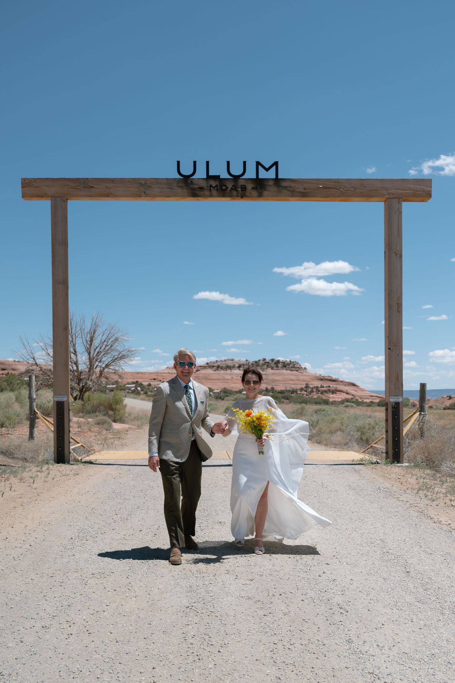 Wedding couple walking hand in hand under rustic ULUM gate in desert.