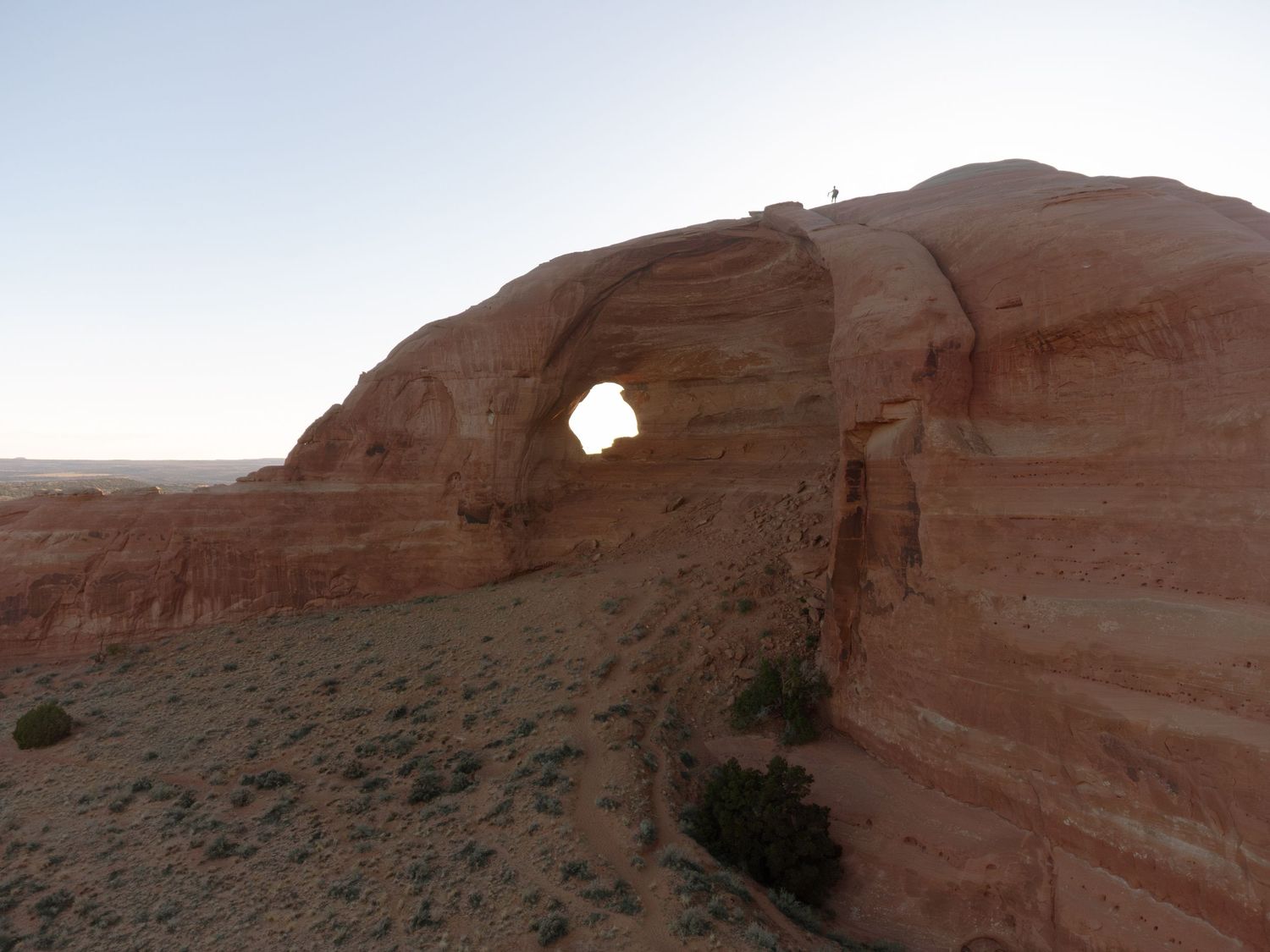 Red sandstone arch formation in an arid desert landscape at dusk, with layered rock walls and sparse desert vegetation below.