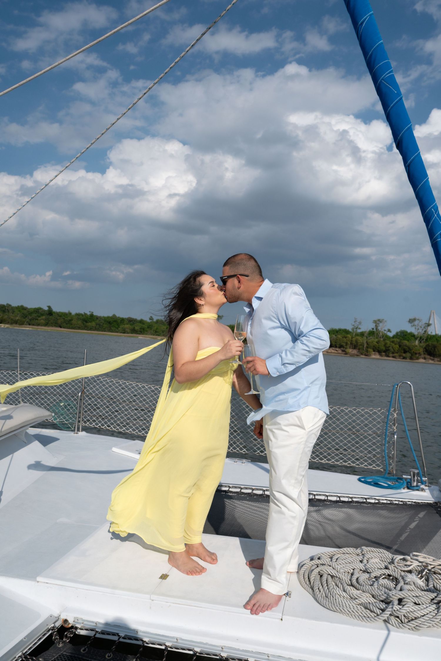 Romantic couple kissing on sailboat deck, woman in yellow dress holding champagne, man in blue shirt, cloudy sky backdrop.