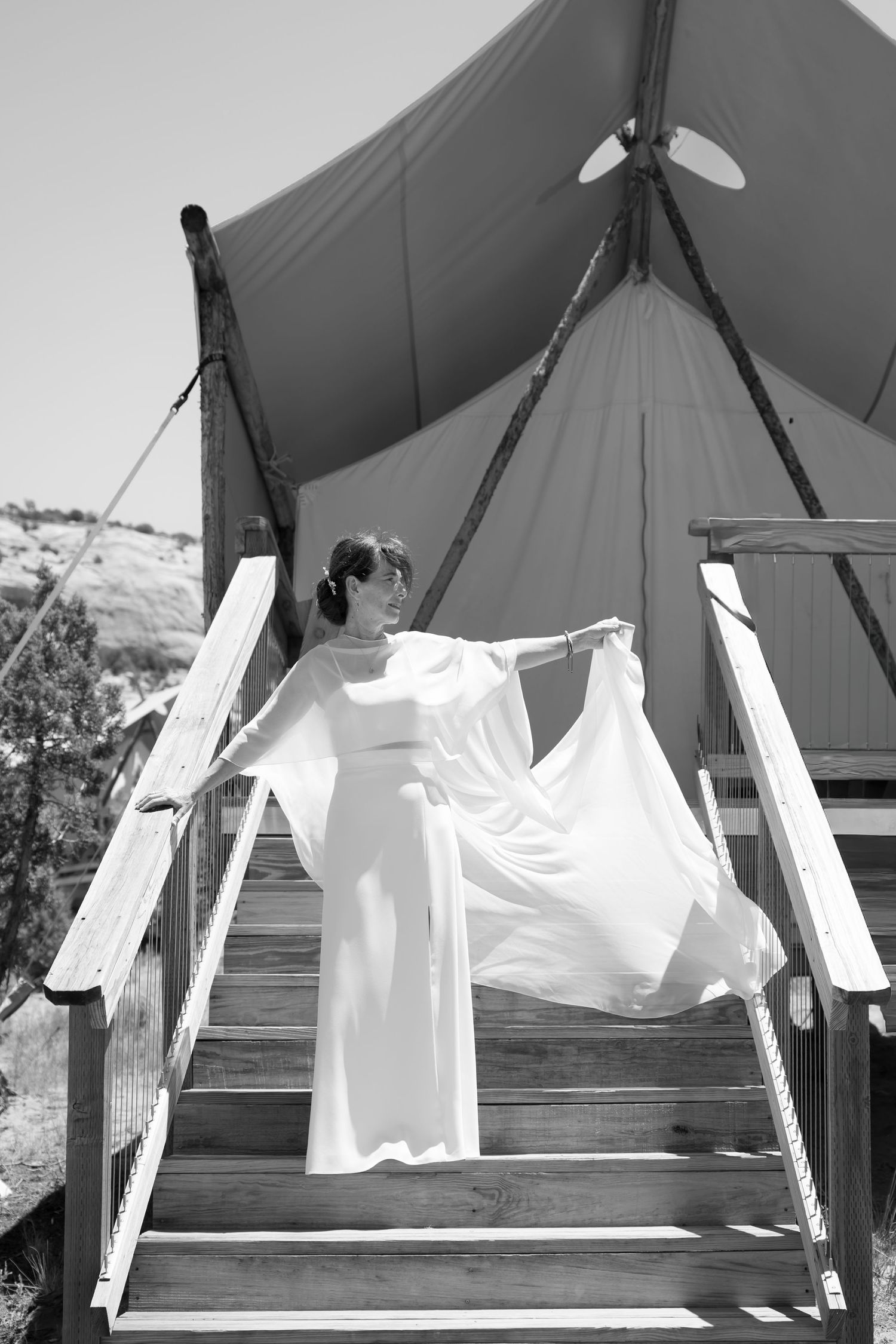 Woman in flowing white gown poses on wooden steps before a canvas glamping tent in black and white photo.