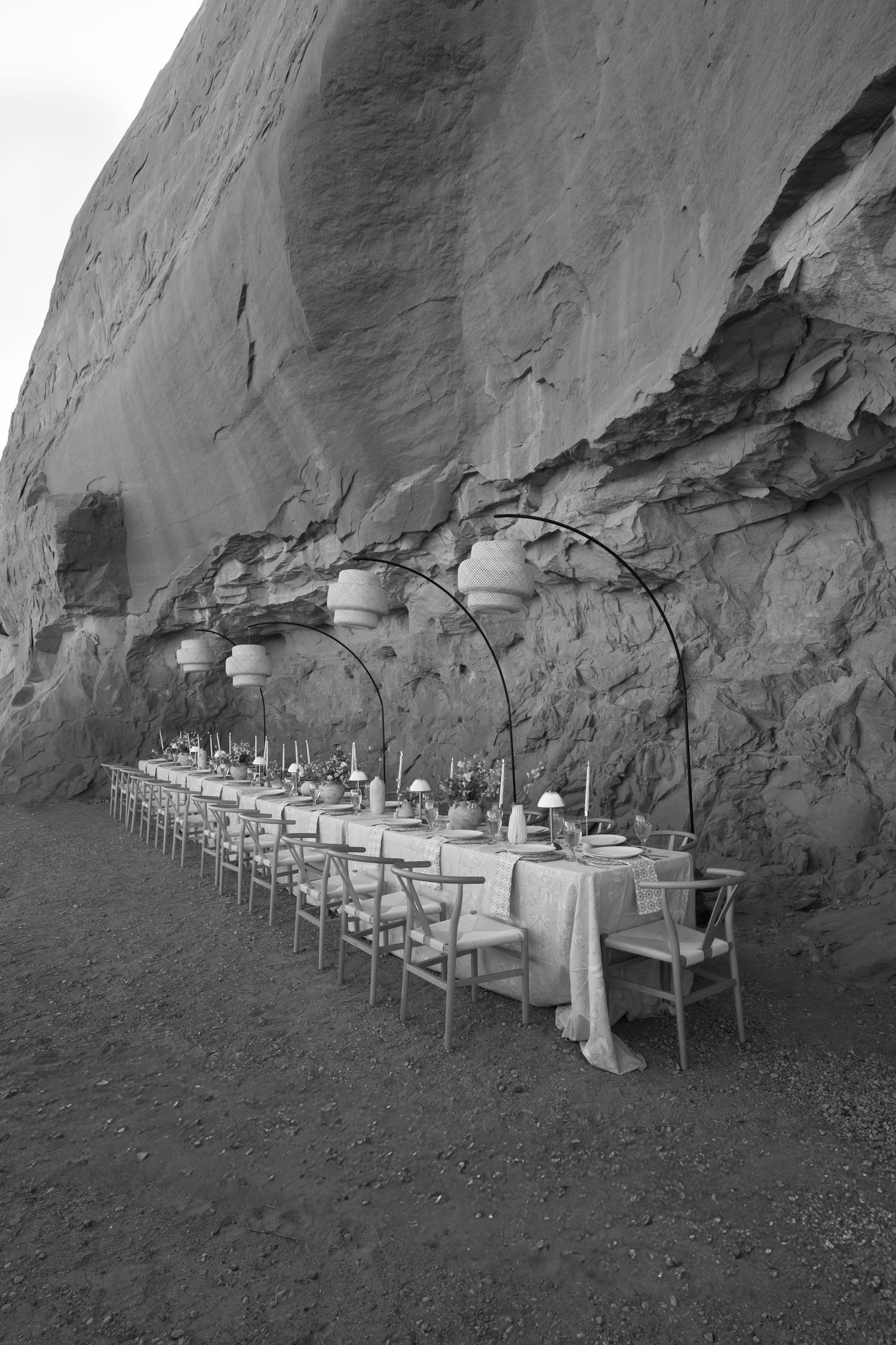 Black and white photo of long elegant wedding reception table set against dramatic red rock canyon wall with hanging lanterns.