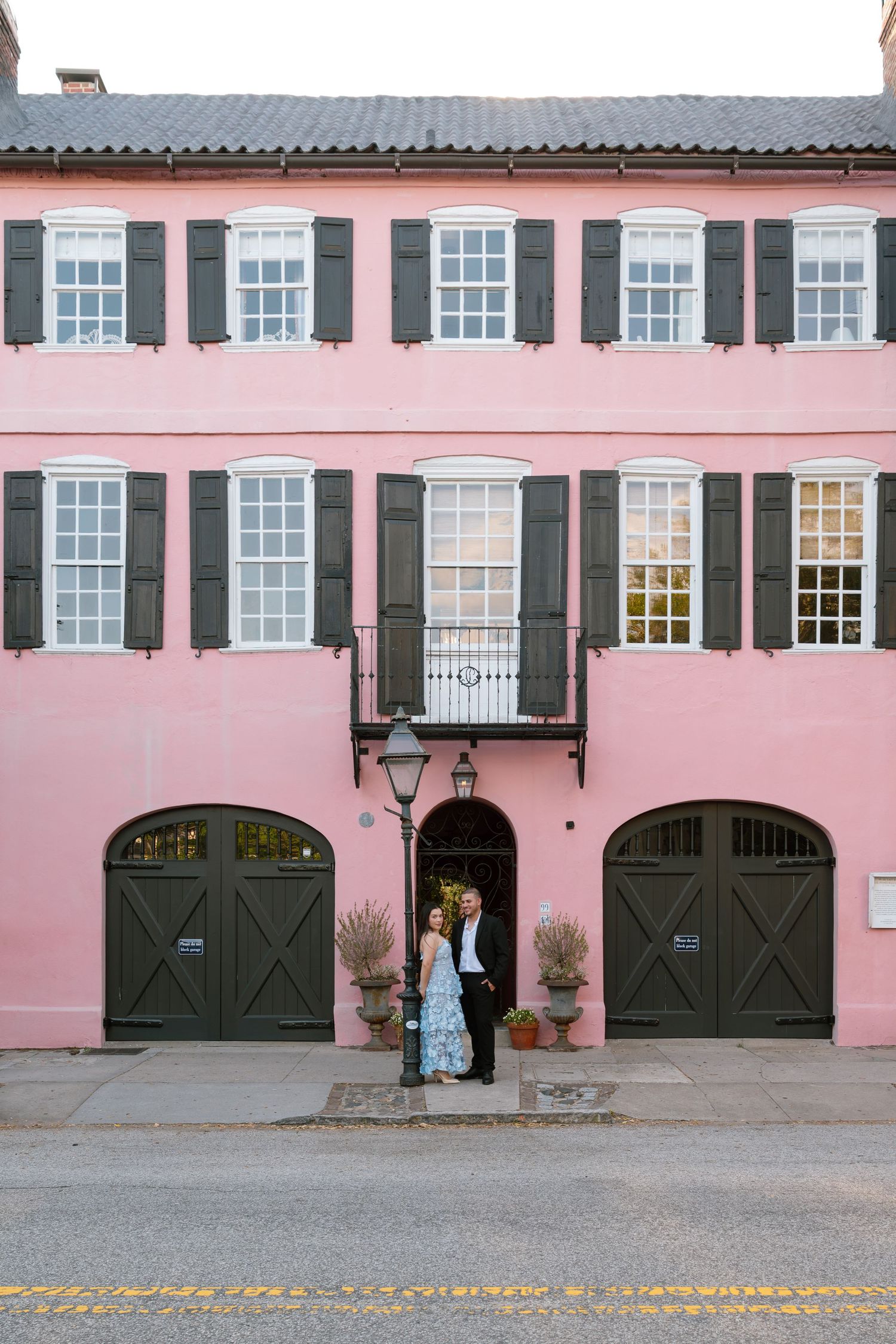 Couple embracing in front of a pink building with dark shutters and arched garage doors in Charleston, SC.