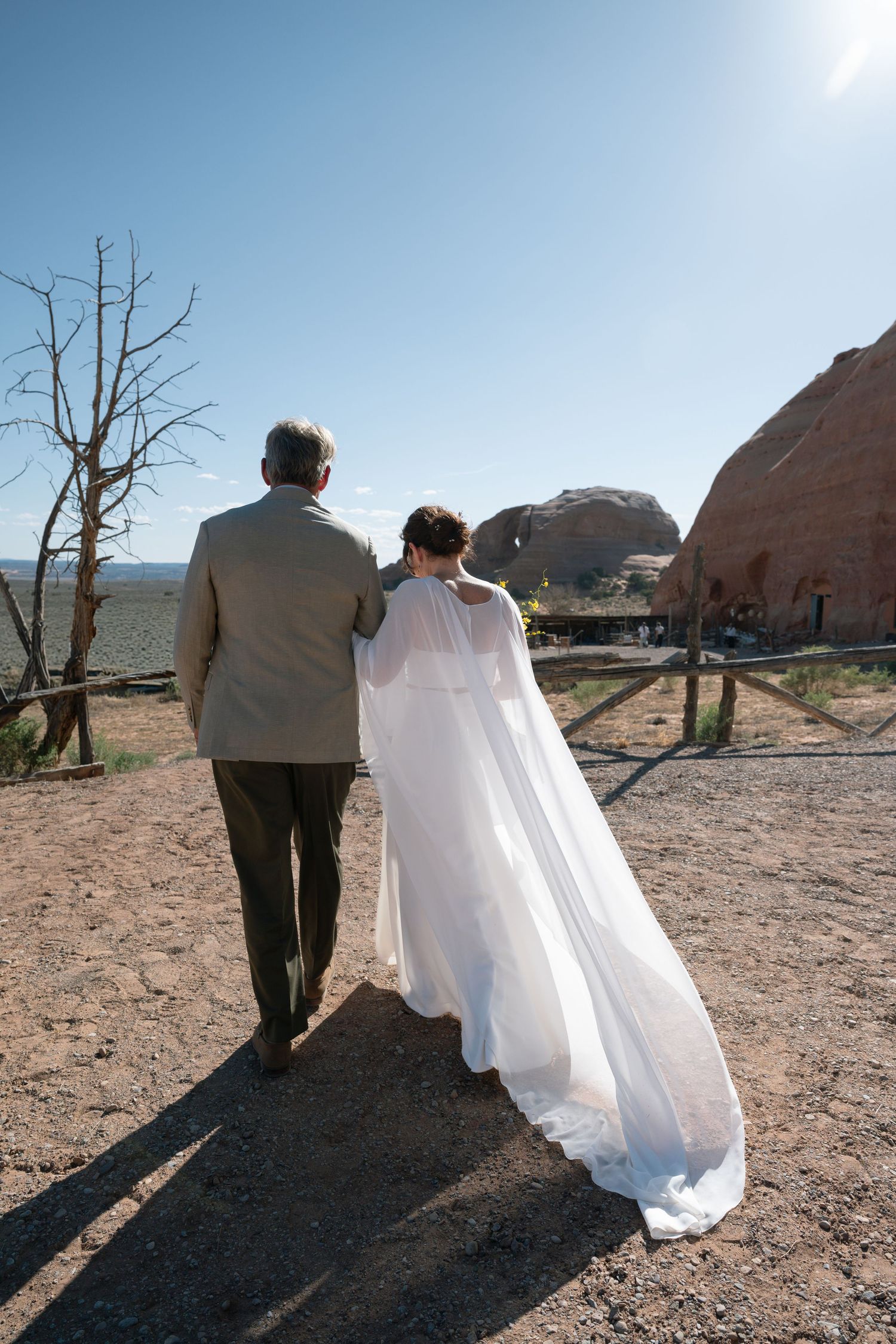 Bride in white cape gown walking with older man toward red rock desert landscape under clear blue sky.