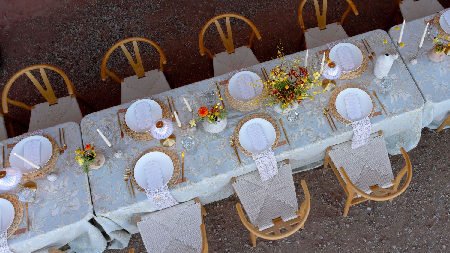 Aerial view of an elegant long dining table set with white plates, gold accents, and floral centerpieces on chairs.