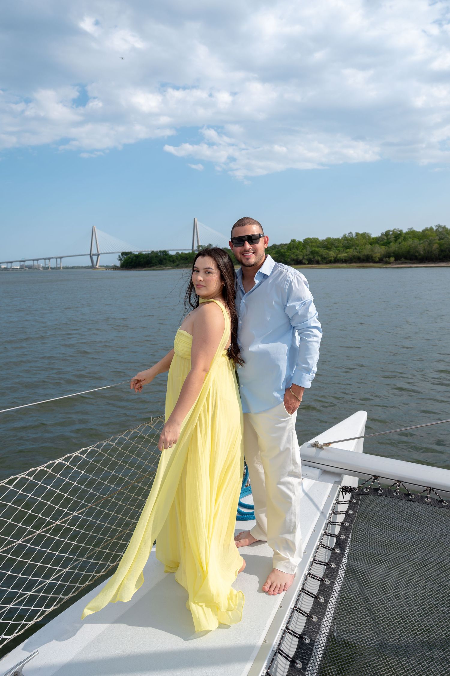 Couple posing on the bow of a sailboat with a suspension bridge and blue sky in the background.