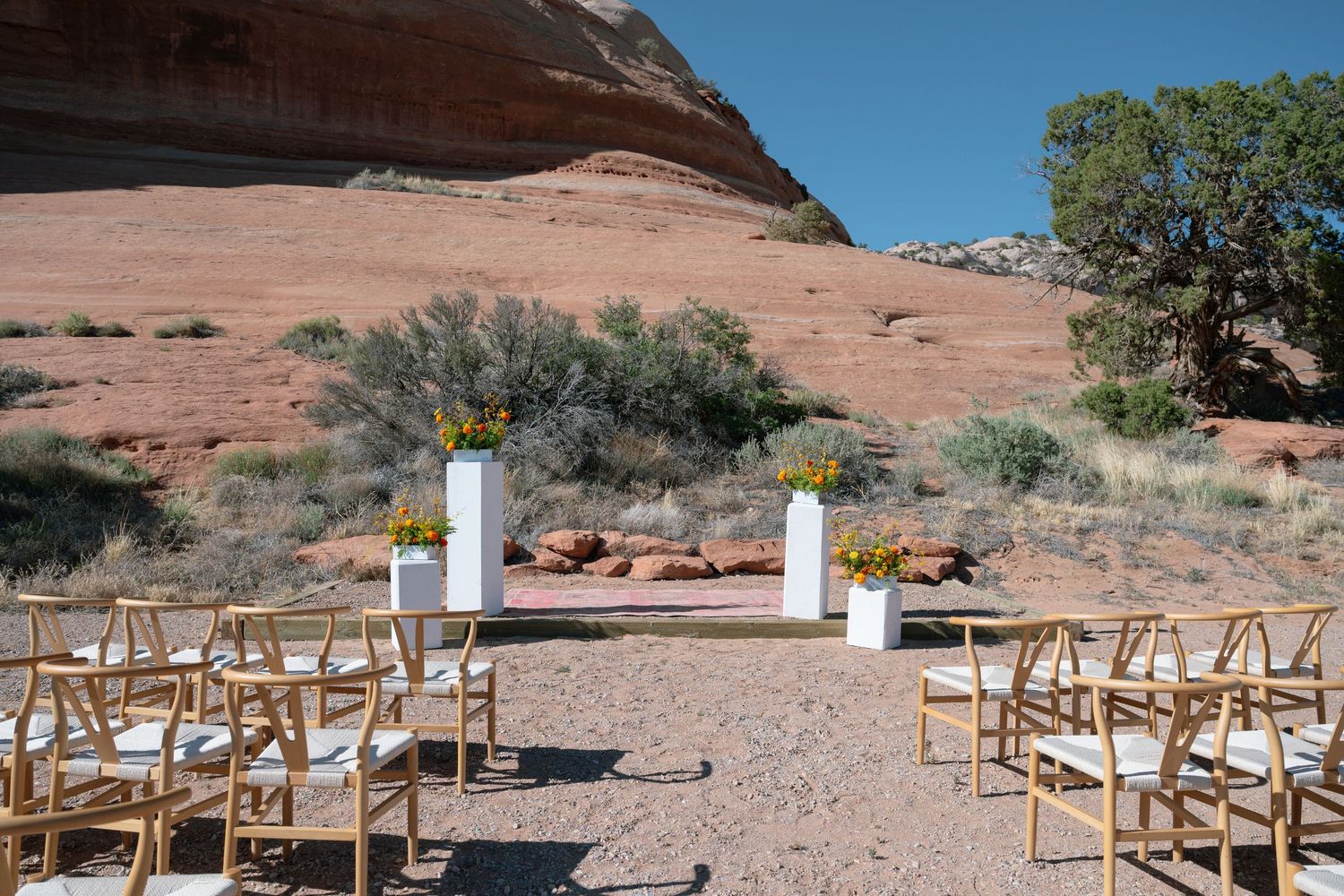 Outdoor desert wedding ceremony setup with wooden chairs and white pedestals against red rock canyon backdrop.