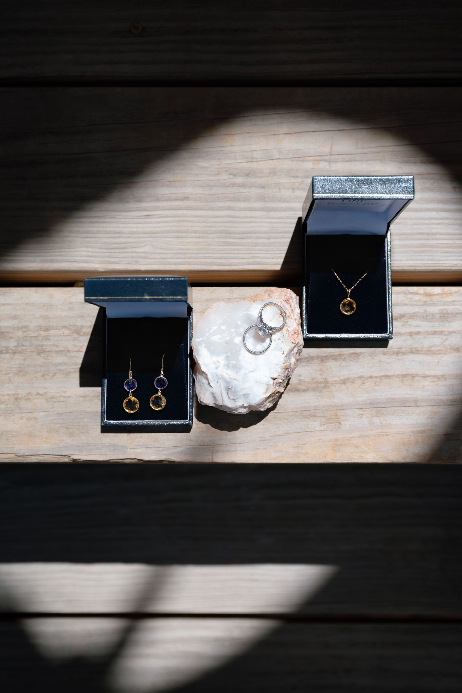 Two open jewelry boxes displaying gold earrings and necklace with gemstones, alongside a ring on a stone, on wooden surface.