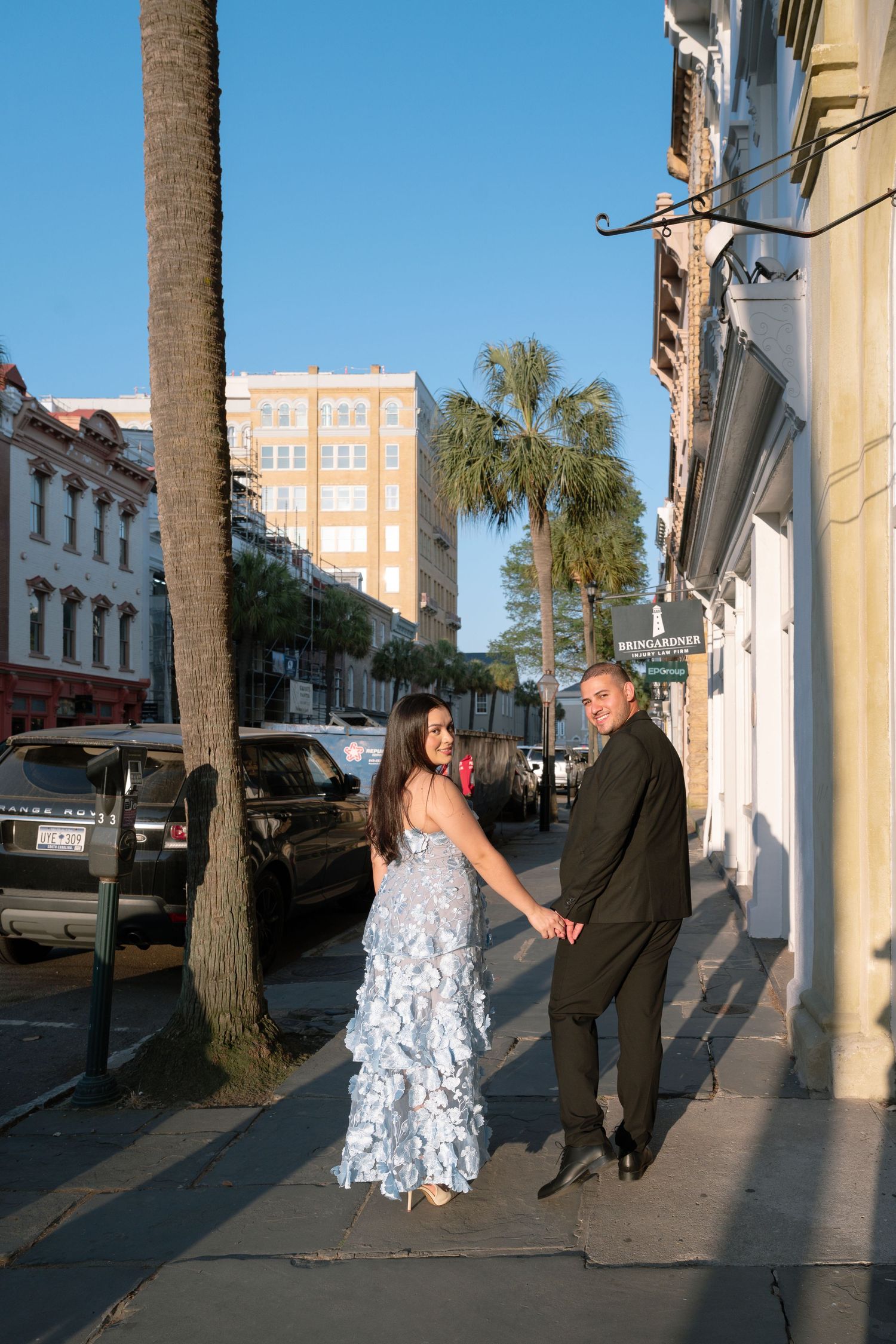 Couple holding hands on a sunny Charleston sidewalk, woman in blue floral gown and man in black suit.