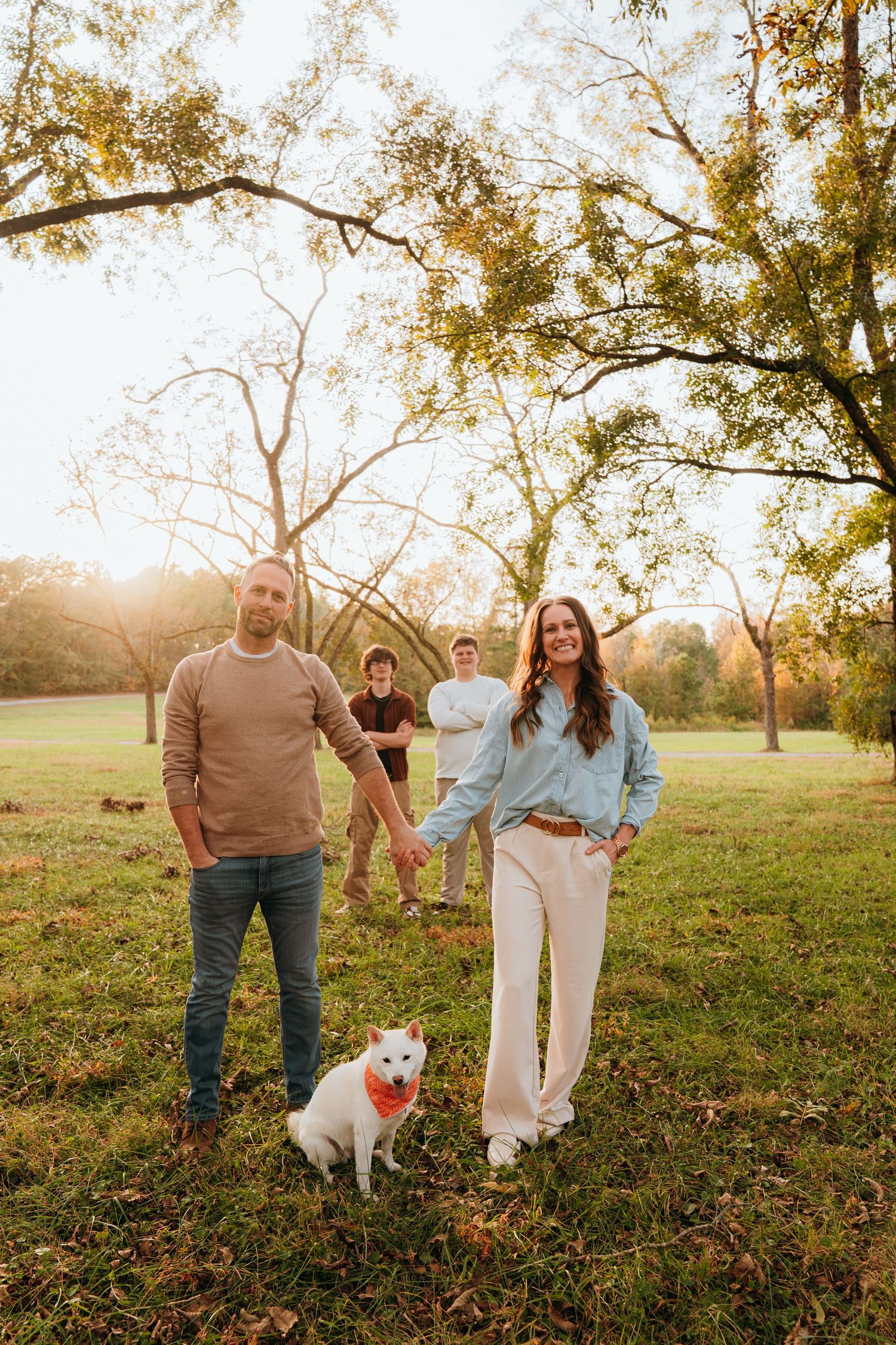 A couple and their small white dog enjoy an autumn sunset in a grassy field with golden light filtering through trees.