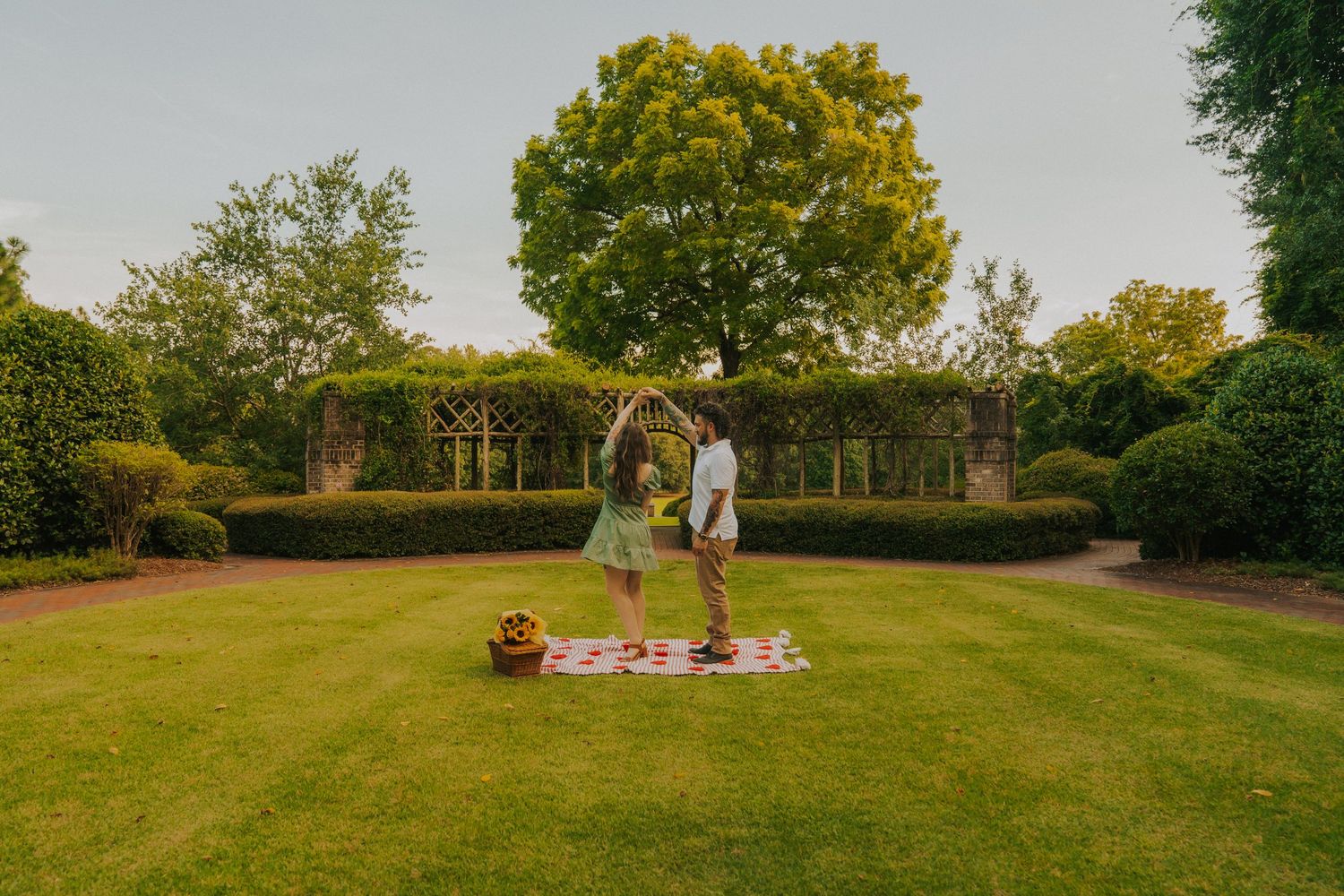 Two people dance together on a small platform in a manicured garden with green archways and trees.