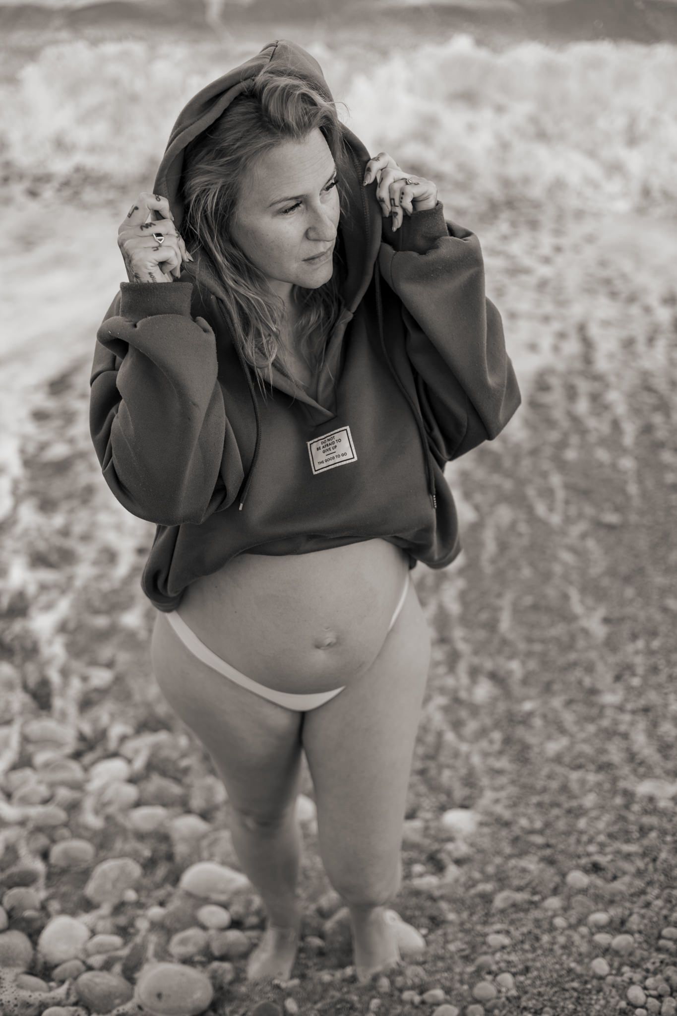 Svartvitt gravidfoto tagen på strand. Kvinna med bar mage och hoodie.