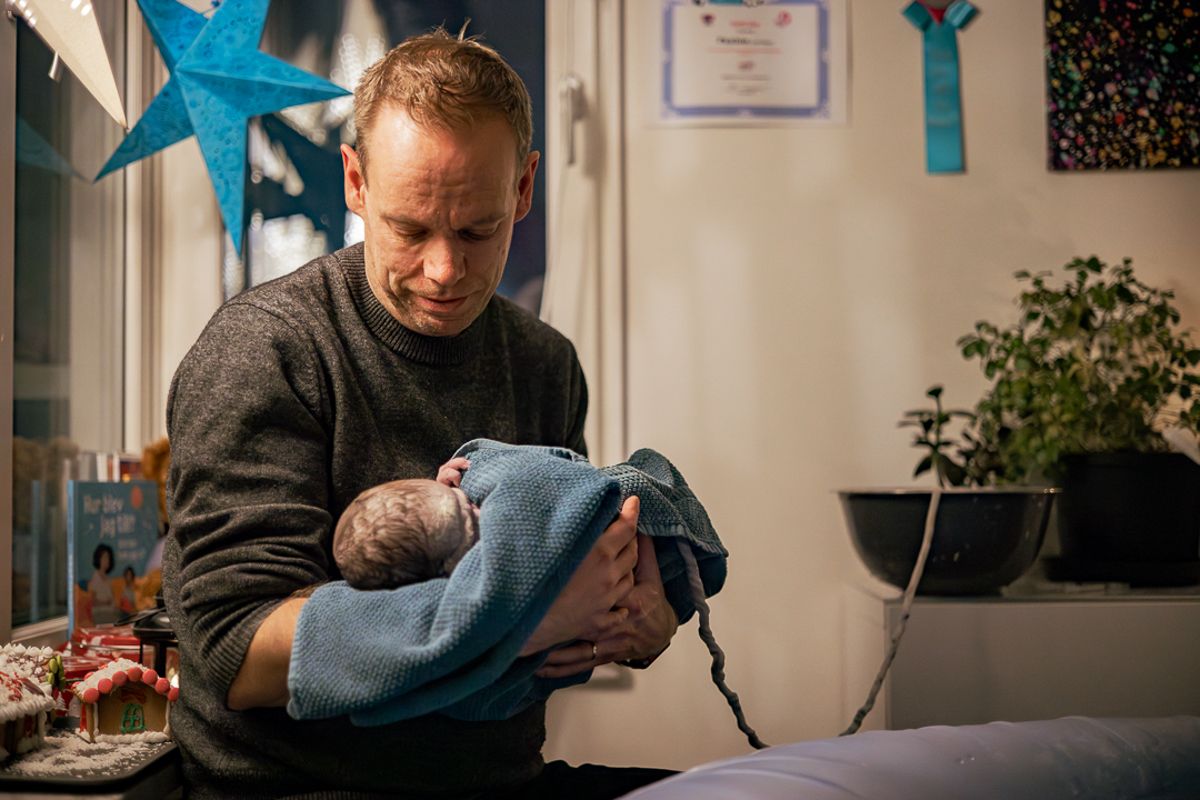 Förlossningsfotografering i Stockholm, hemfödsel. Pappa sitter med nyfödd bebis i famnen. Julstjärnan lyser. Navelsträngen går till en skål