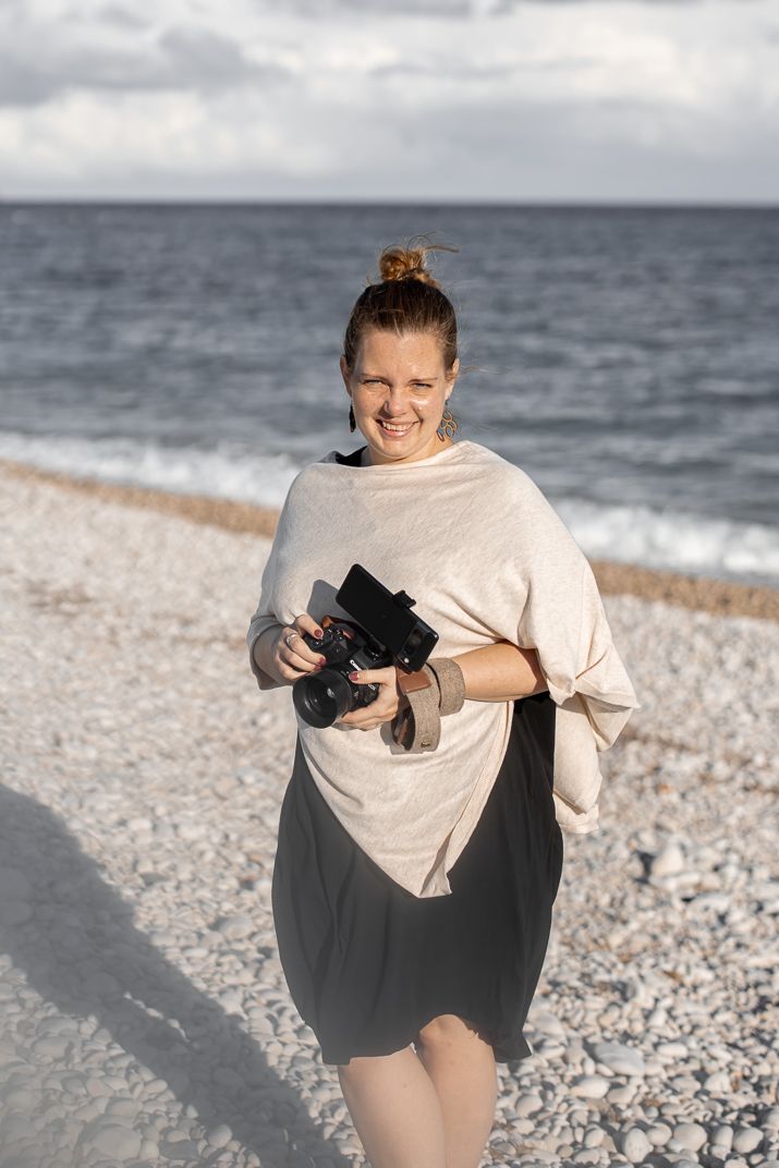 A photographer in a cream sweater and dark skirt holding a camera stands on a pebble beach by the ocean.