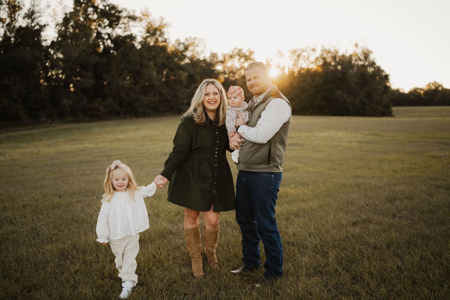 Family of four posing in a sunlit green field at golden hour, parents holding children with trees in the background.