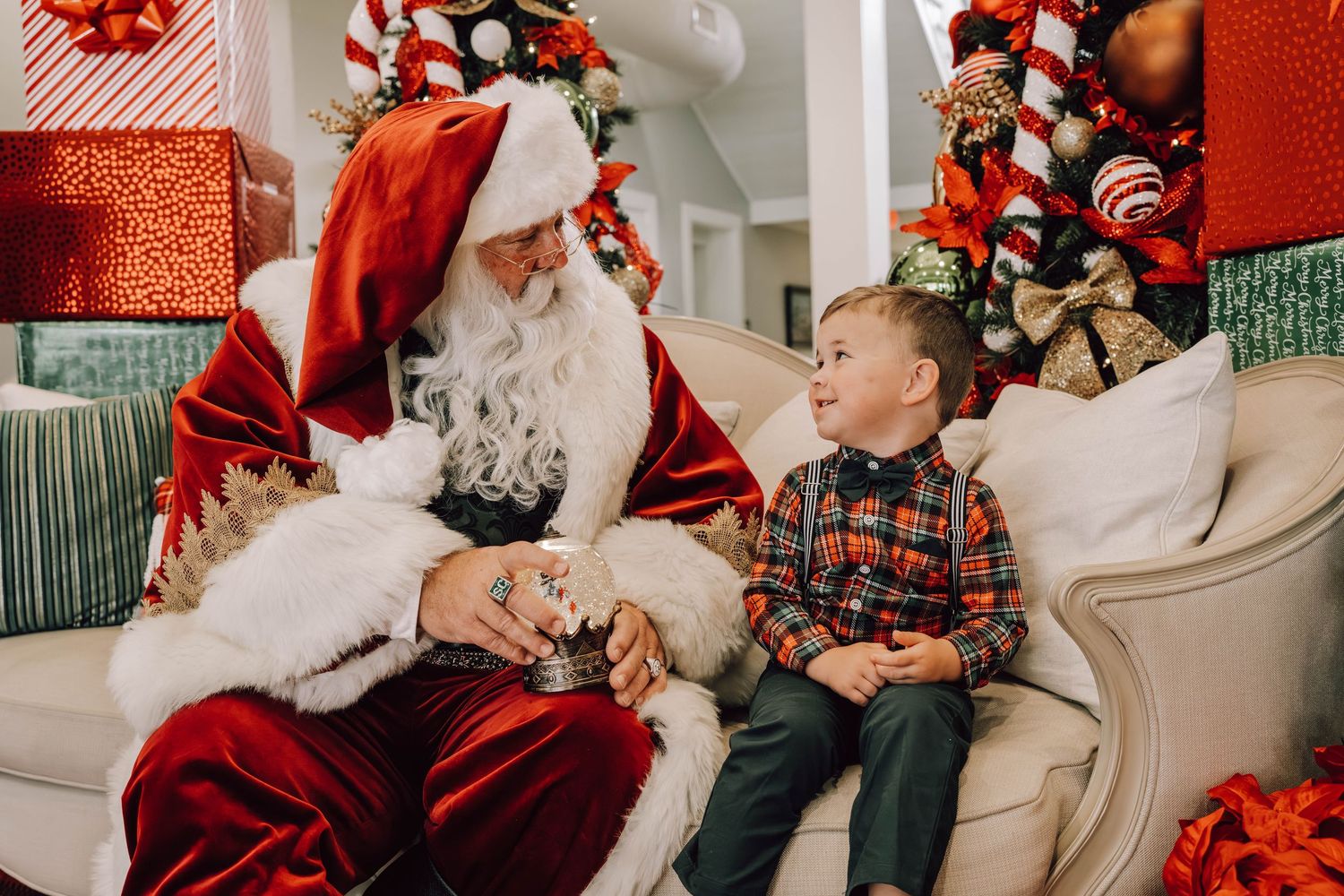 Santa Claus sitting on a sofa with a young boy near a decorated Christmas tree with gifts.