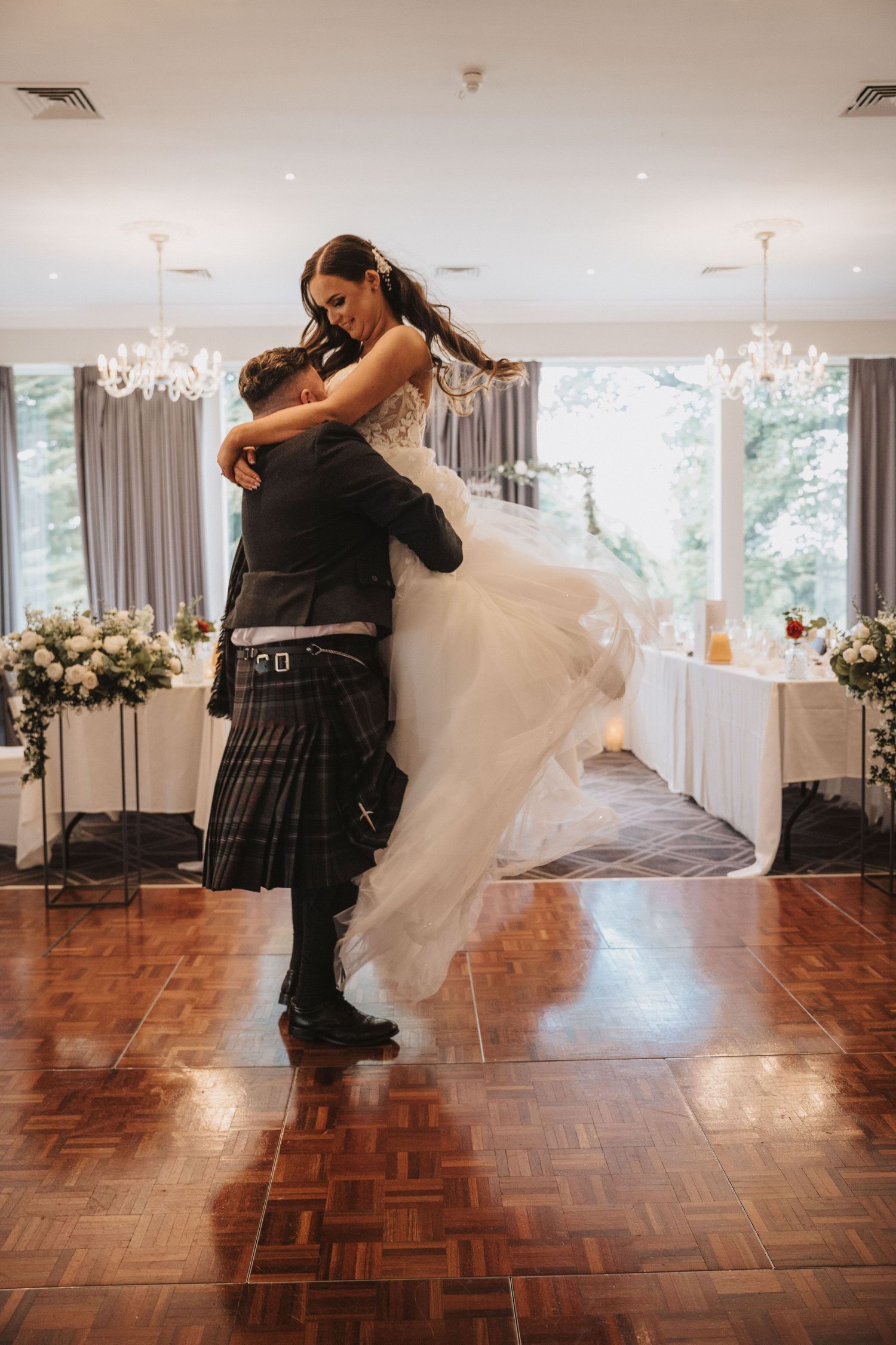 A romantic wedding dance moment captured on a parquet floor with elegant floral decorations in the background.