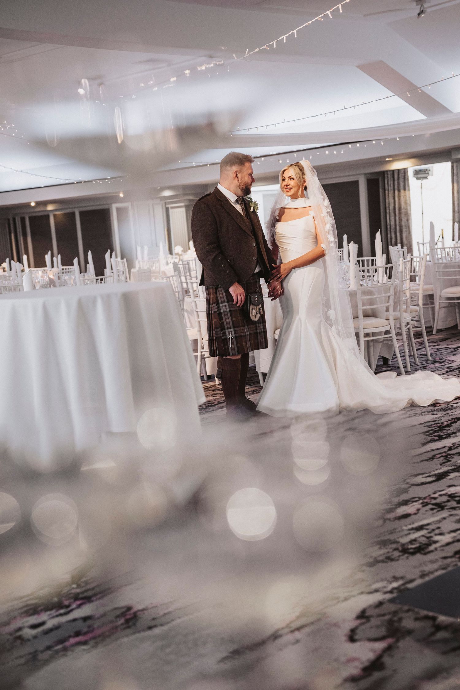 A couple shares a romantic moment in a modern white wedding venue with bokeh light effects in the foreground.