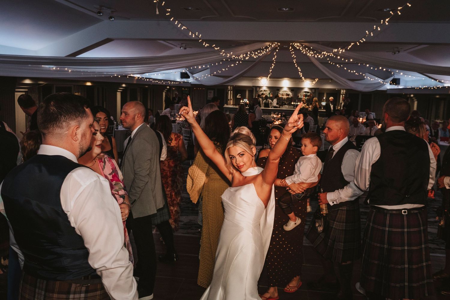 Wedding guests dance and celebrate on a dimly lit dance floor decorated with string lights during an evening reception.