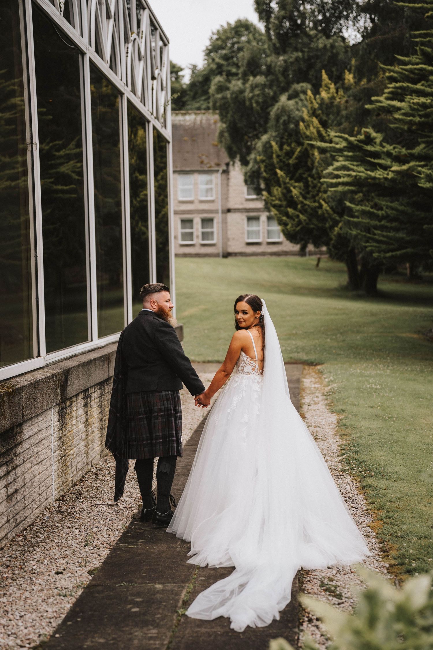 Couple in formal wedding attire walking along a brick building with long windows and green lawn in the background.