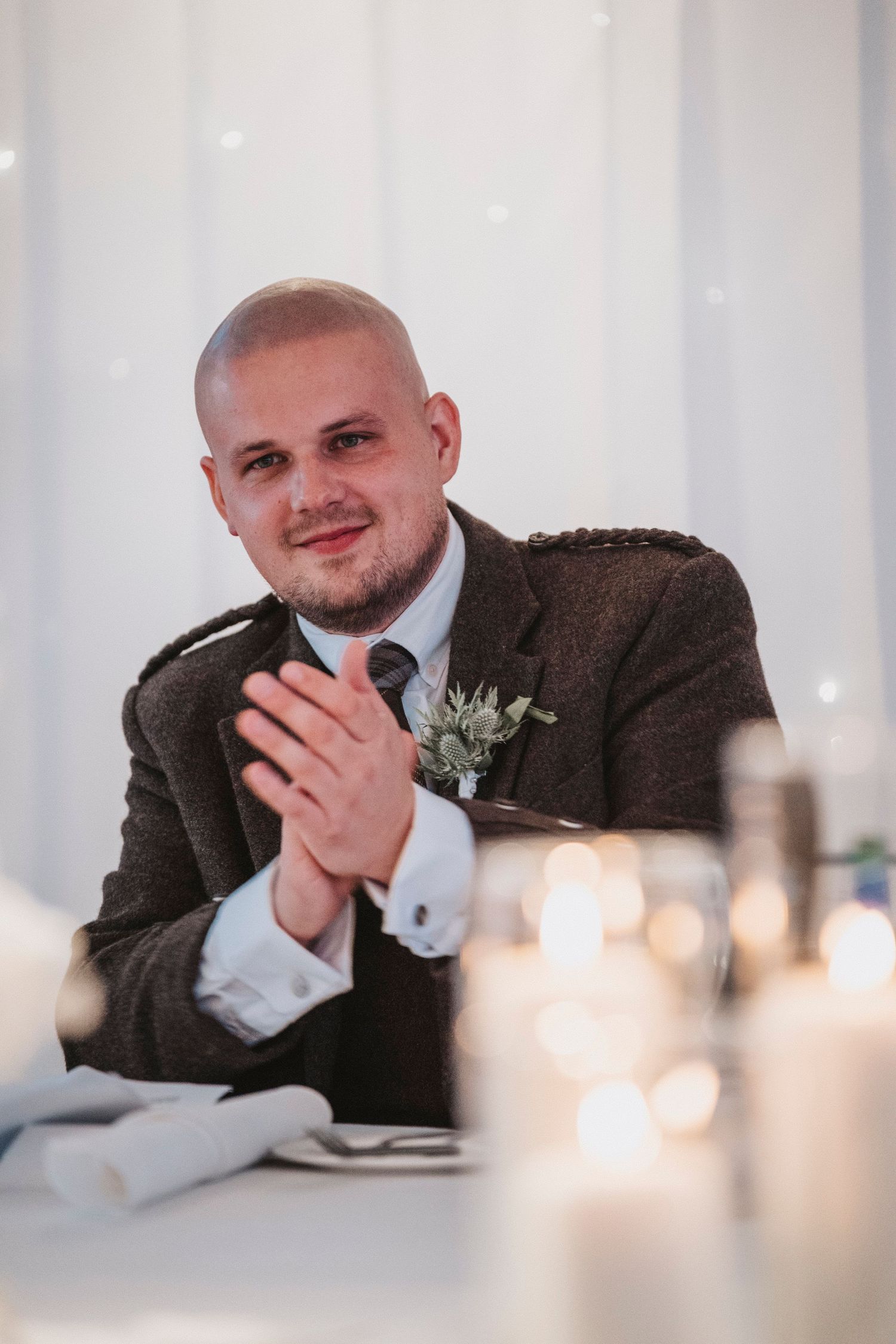Four candid wedding photos show a guest in a brown suit resting their chin on their hand at a table with glowing candles.