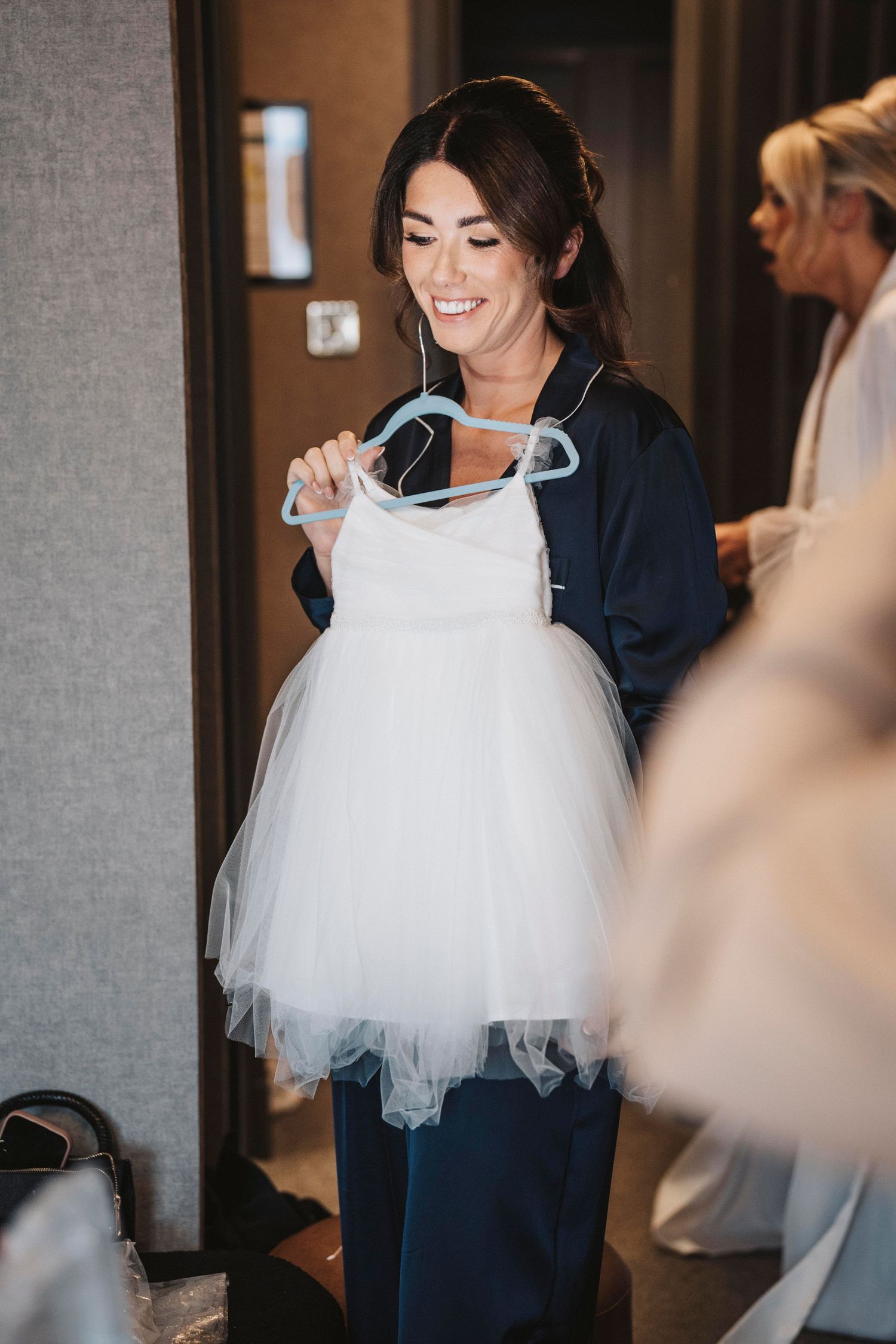 A person wearing a navy blue dress holds up a white tulle wedding dress on a hanger in an indoor setting.