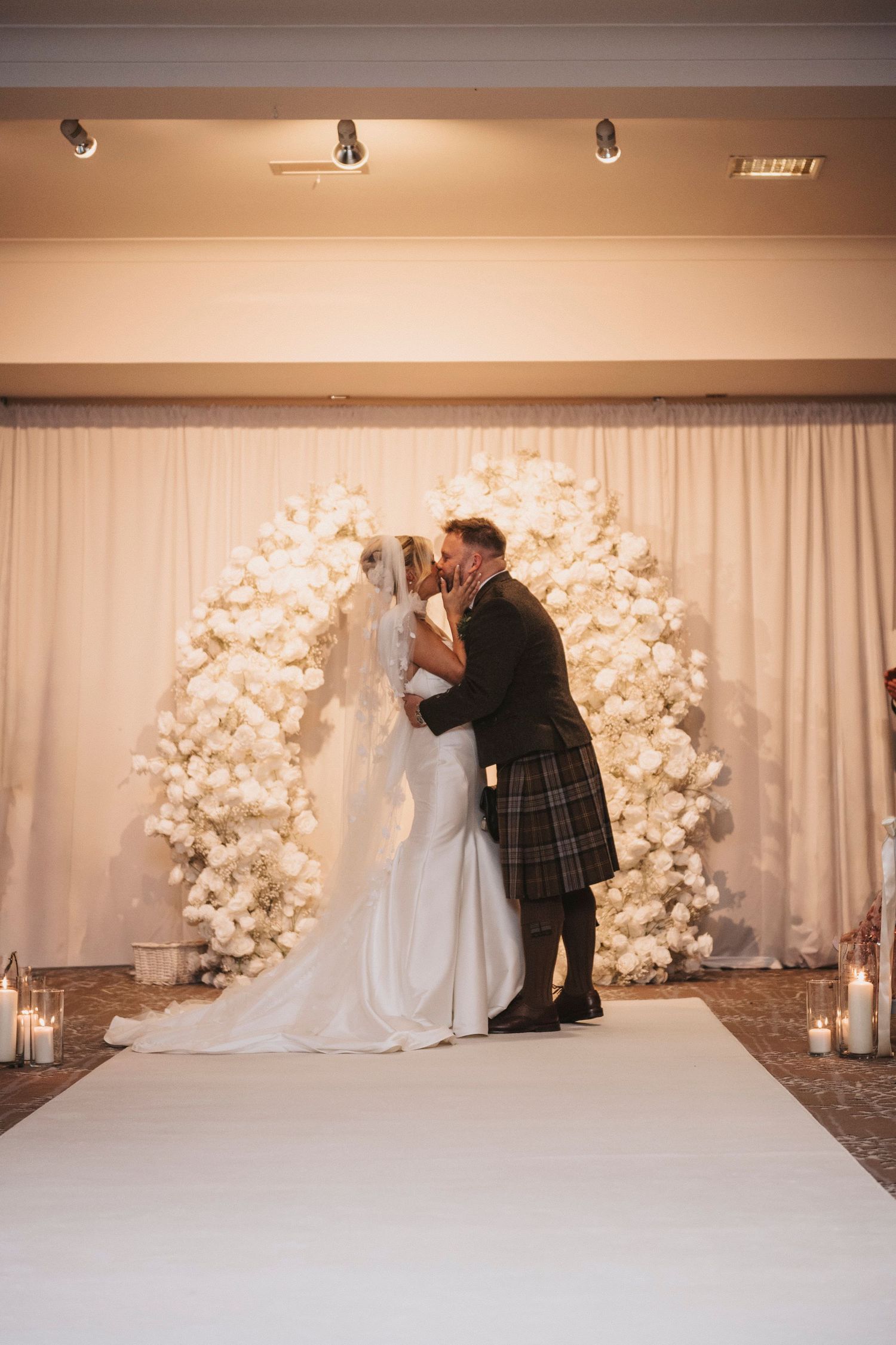 Wedding photographer adjusts bride's dress train against elegant white floral backdrop with glowing candles.