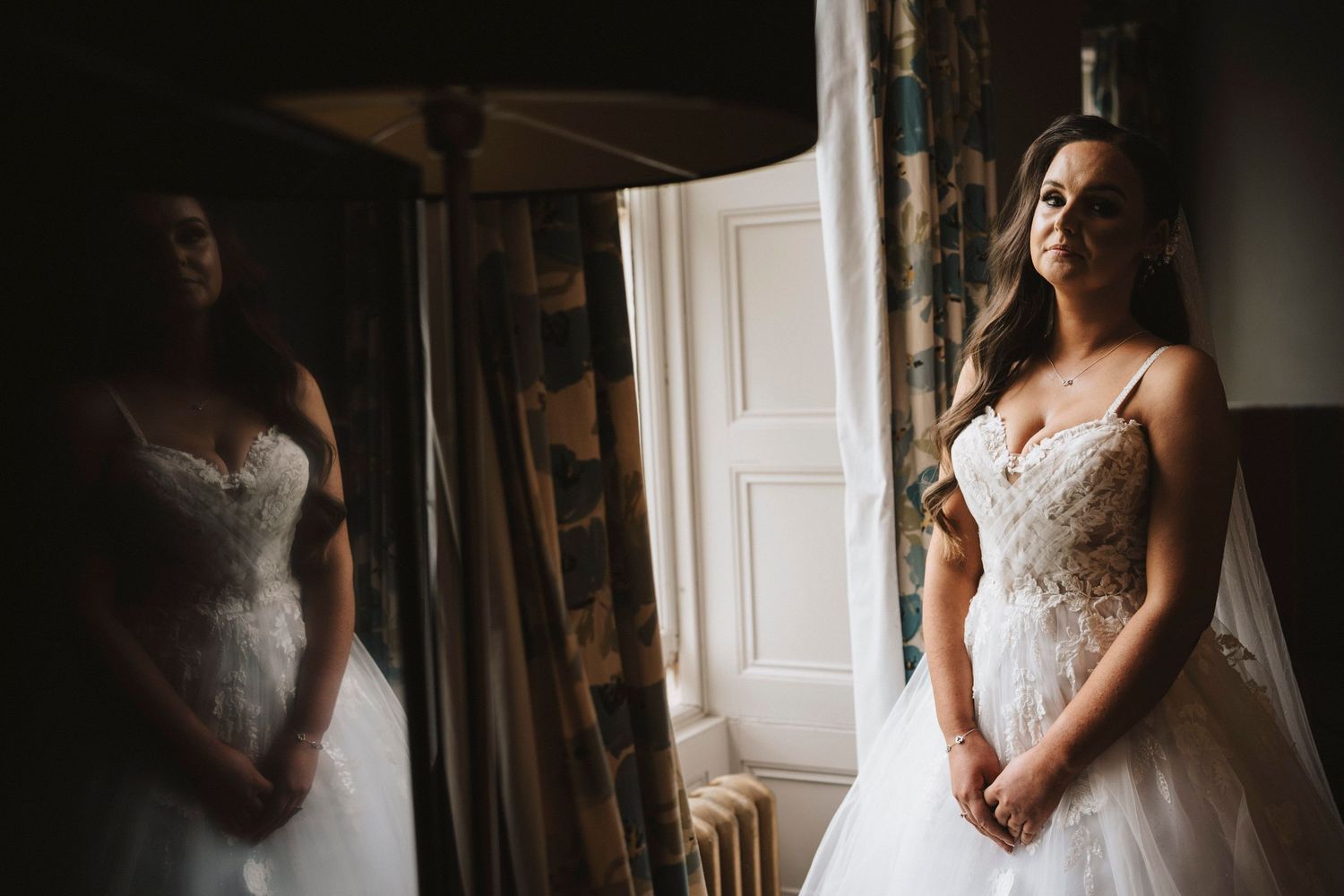 A bride in a white lace wedding gown stands in soft natural light near a mirror and white door.