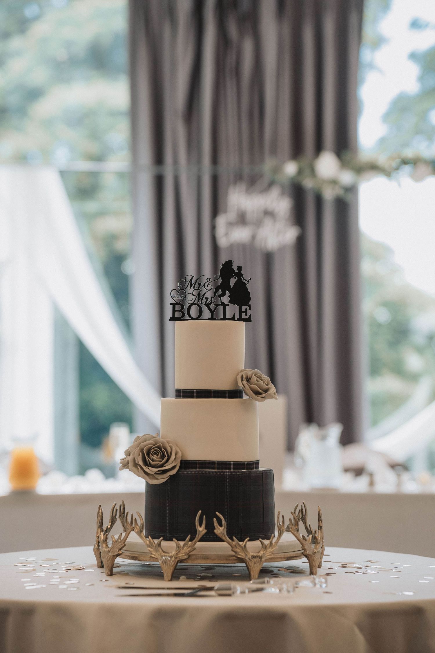 Black and white modern tiered wedding cake with Love topper against grey curtains.