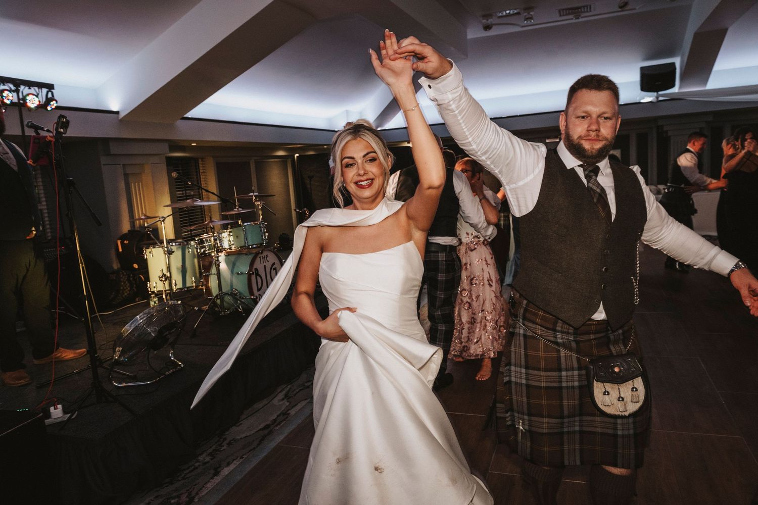 A bride and groom dancing joyfully at their wedding reception with a band setup visible in the background.