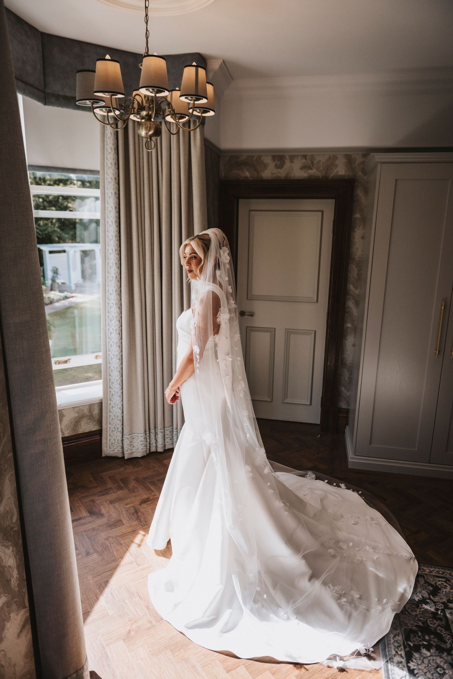 A bride in an elegant white wedding gown with long train and veil stands by a window in a vintage-style room.