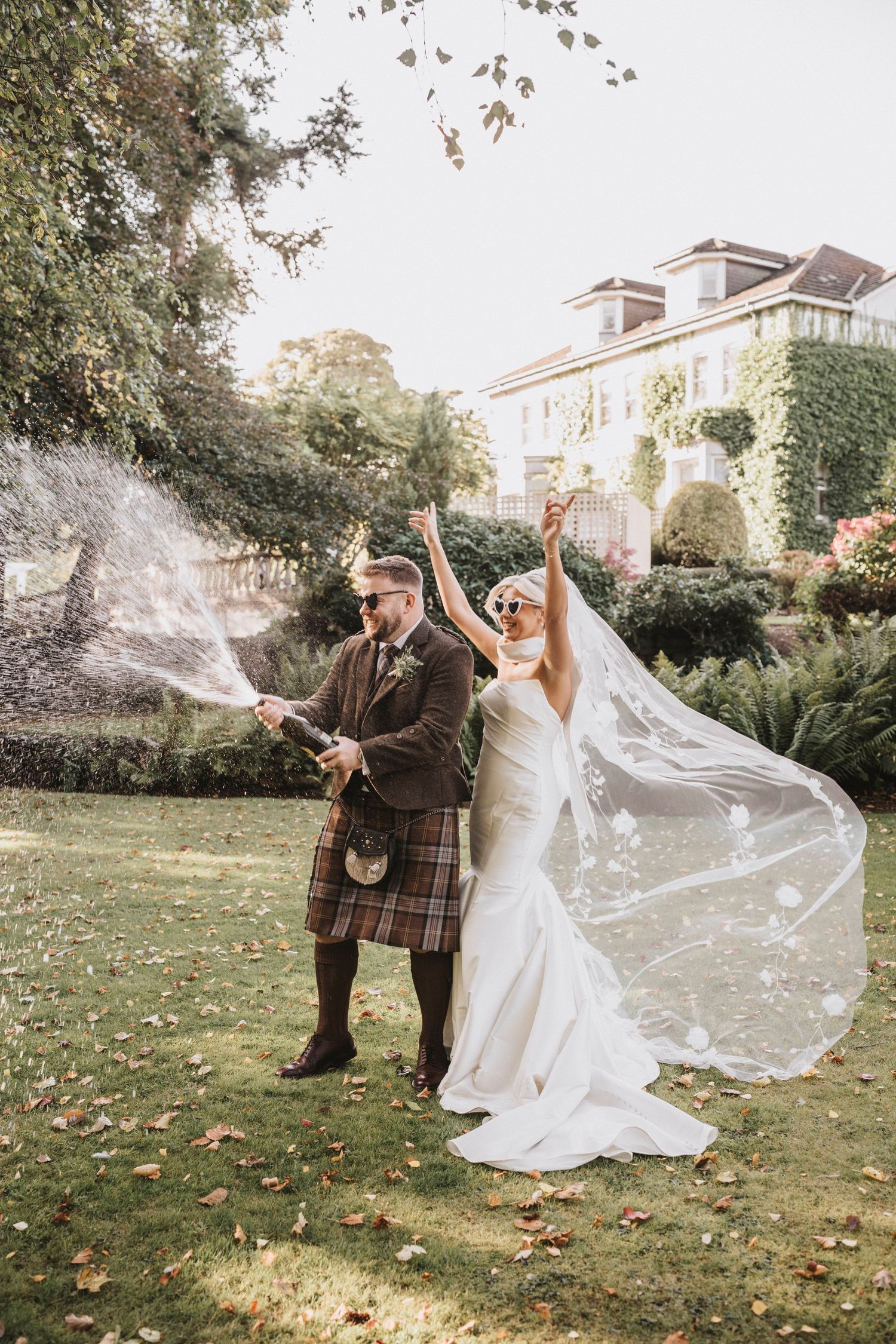 Wedding couple celebrates outdoors with flowing veil and traditional Scottish attire in autumn garden setting.