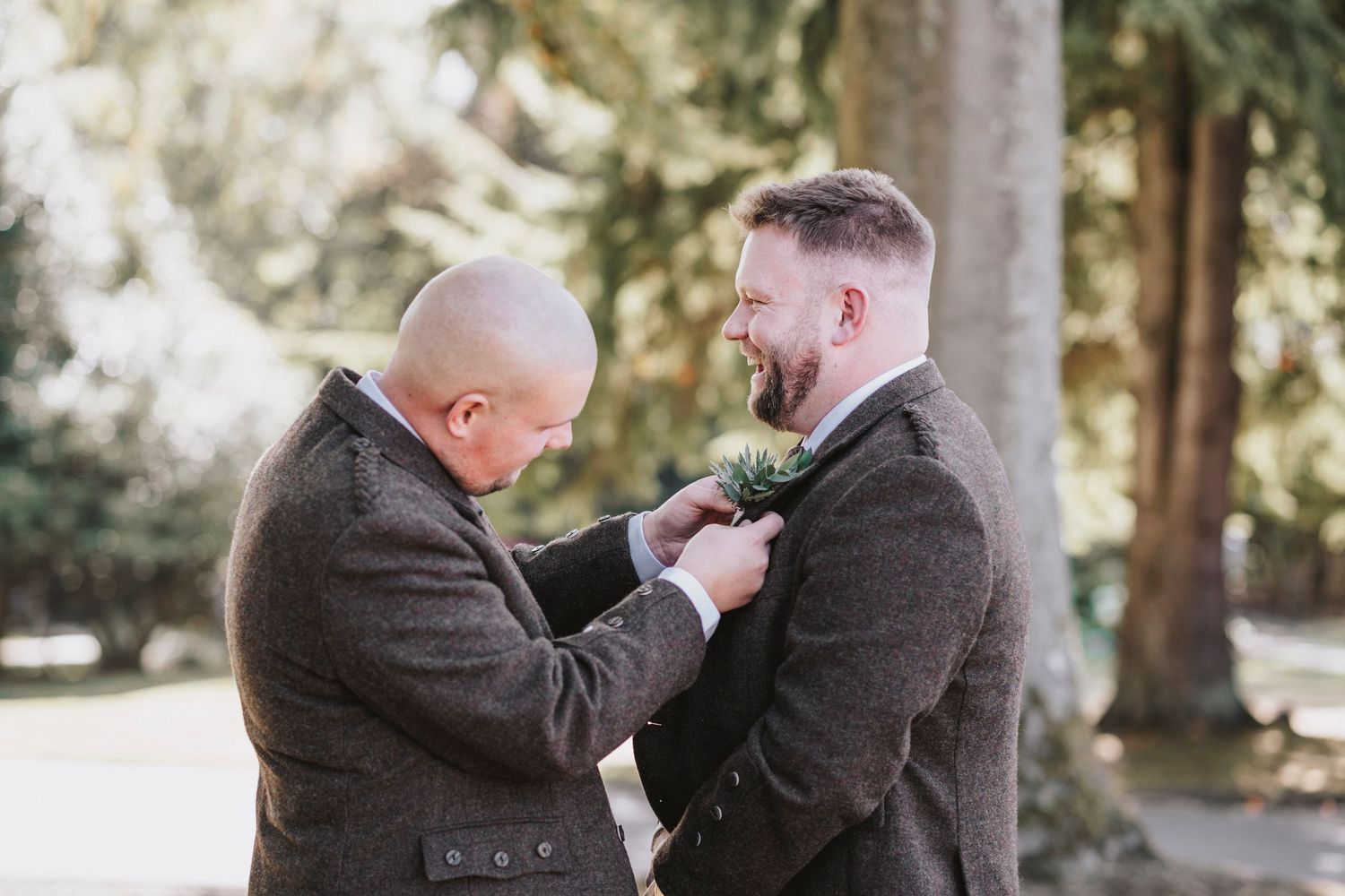 Two people in dark suits share a tender moment while adjusting a boutonniere in a sunlit outdoor setting.