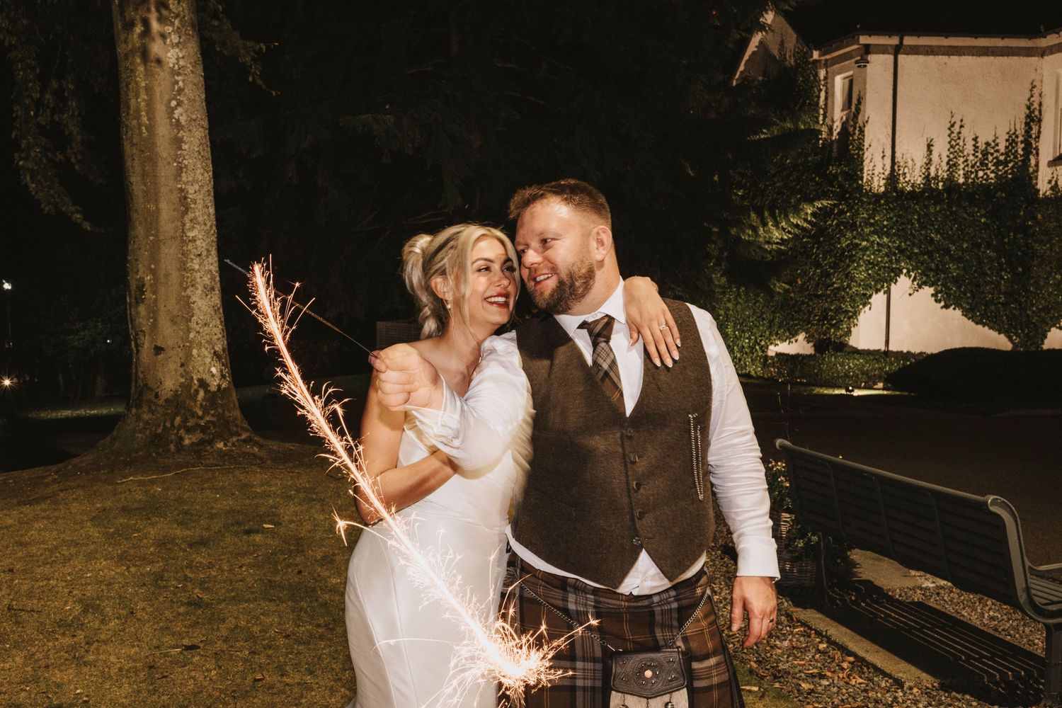 A couple celebrates their wedding night outdoors holding sparklers in a romantic nighttime setting.