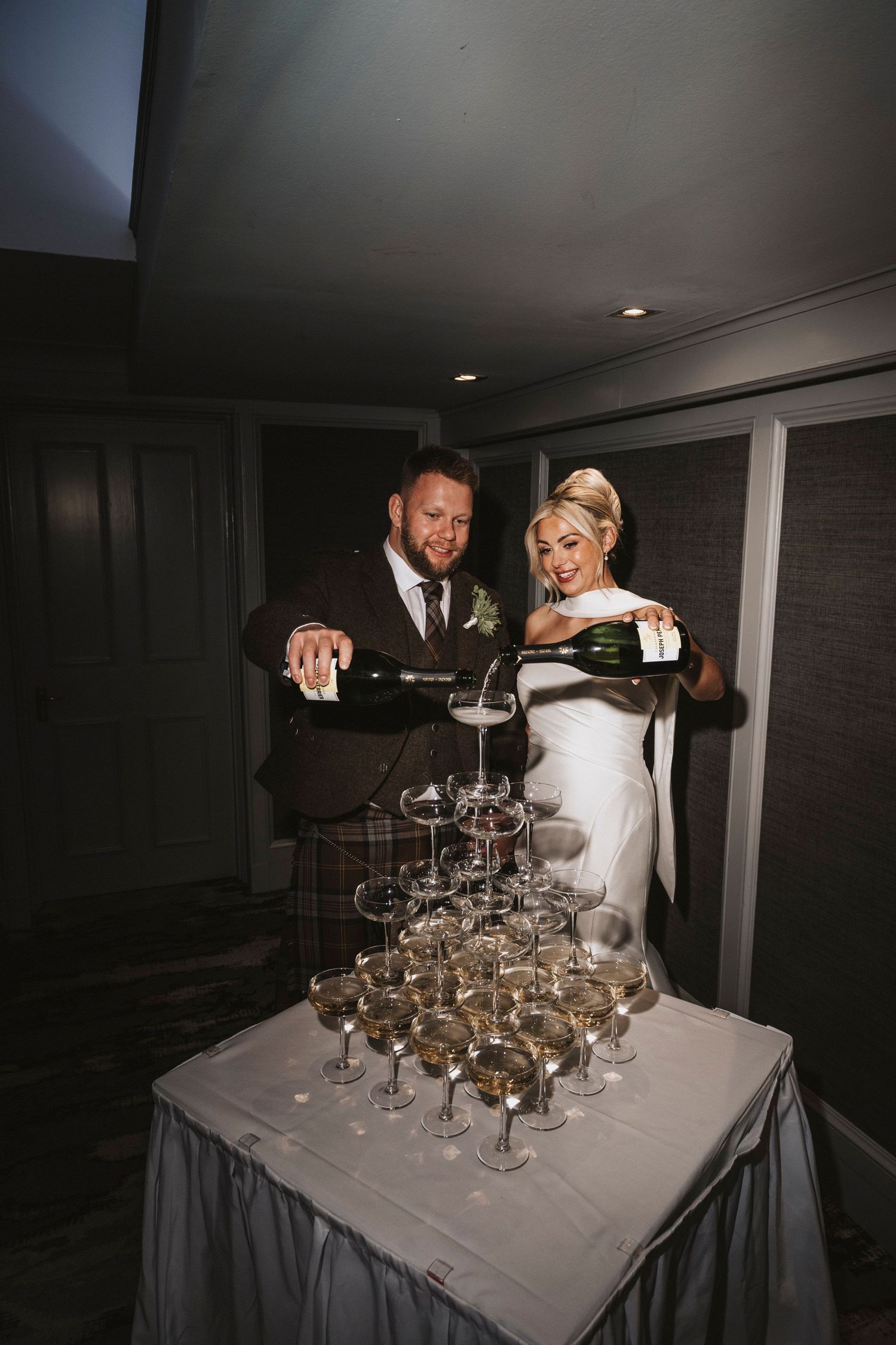 A couple pours champagne into a pyramid of glasses stacked on a table at a wedding reception.