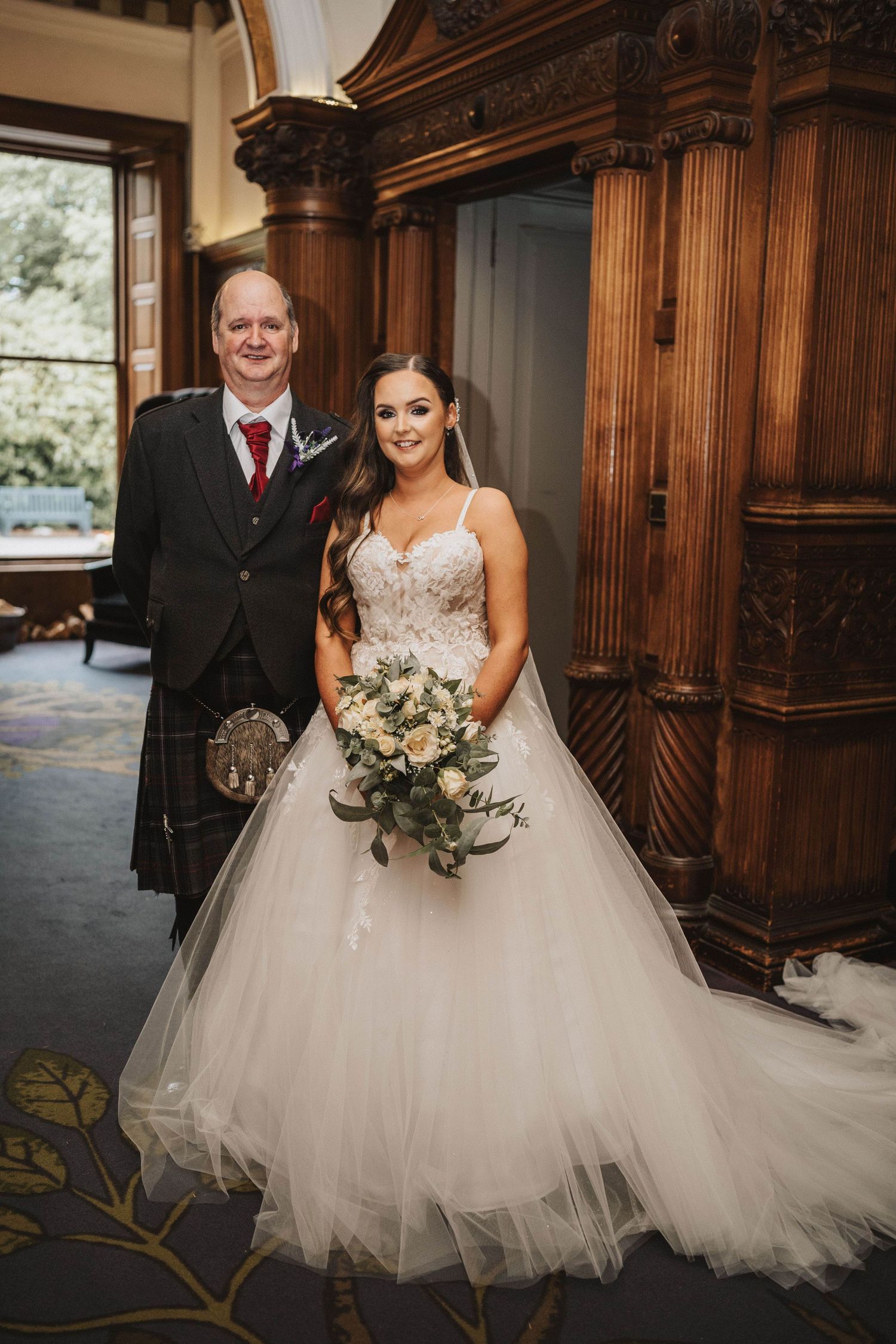 A Scottish wedding couple poses for portraits in front of ornate wooden panels at their traditional celebration.